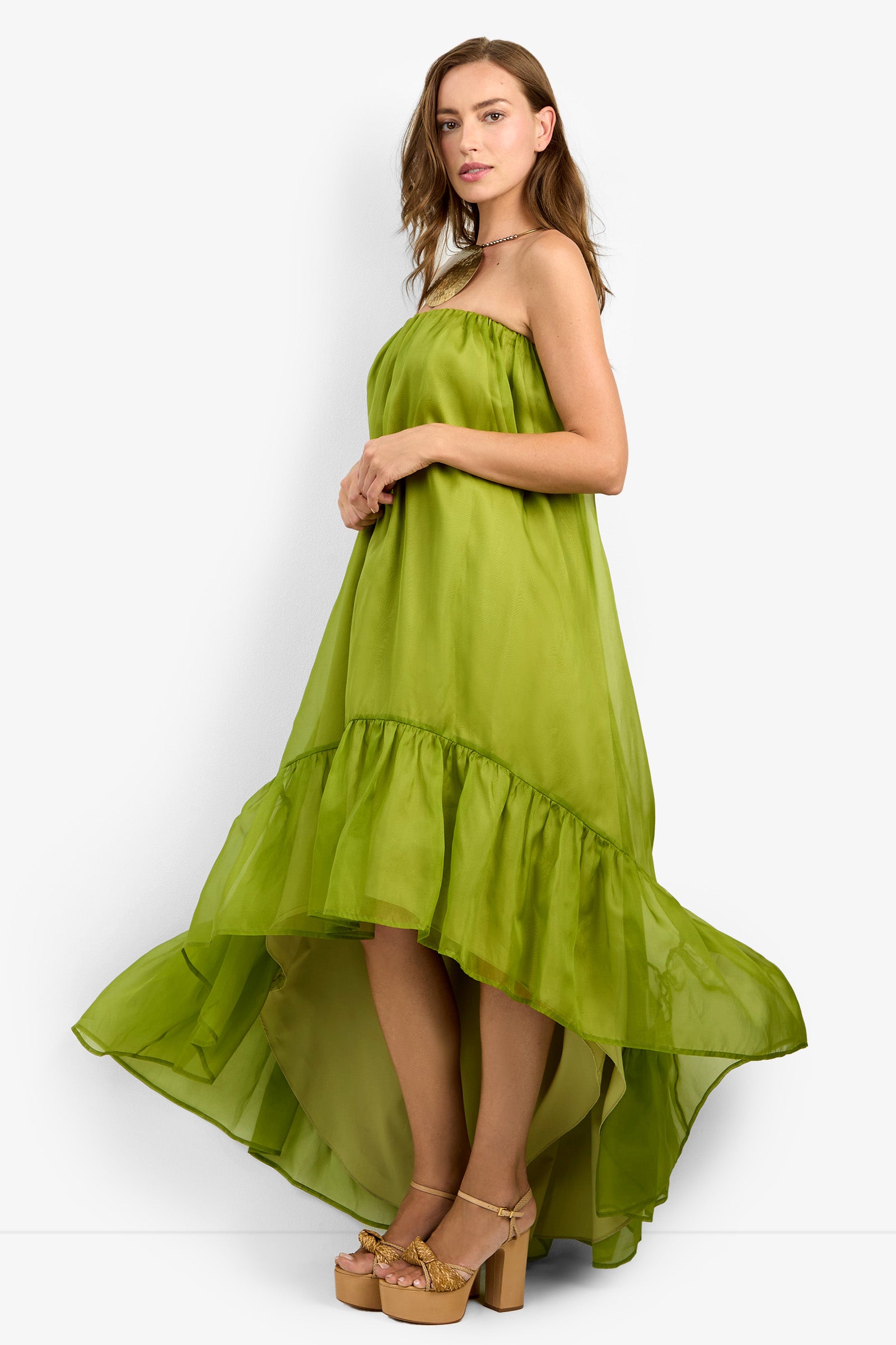 A woman stands sideways, looking at the camera against a plain white background, wearing the ARIAH DRESS—a flowing, strapless, lime green high-low dress—paired with platform sandals.