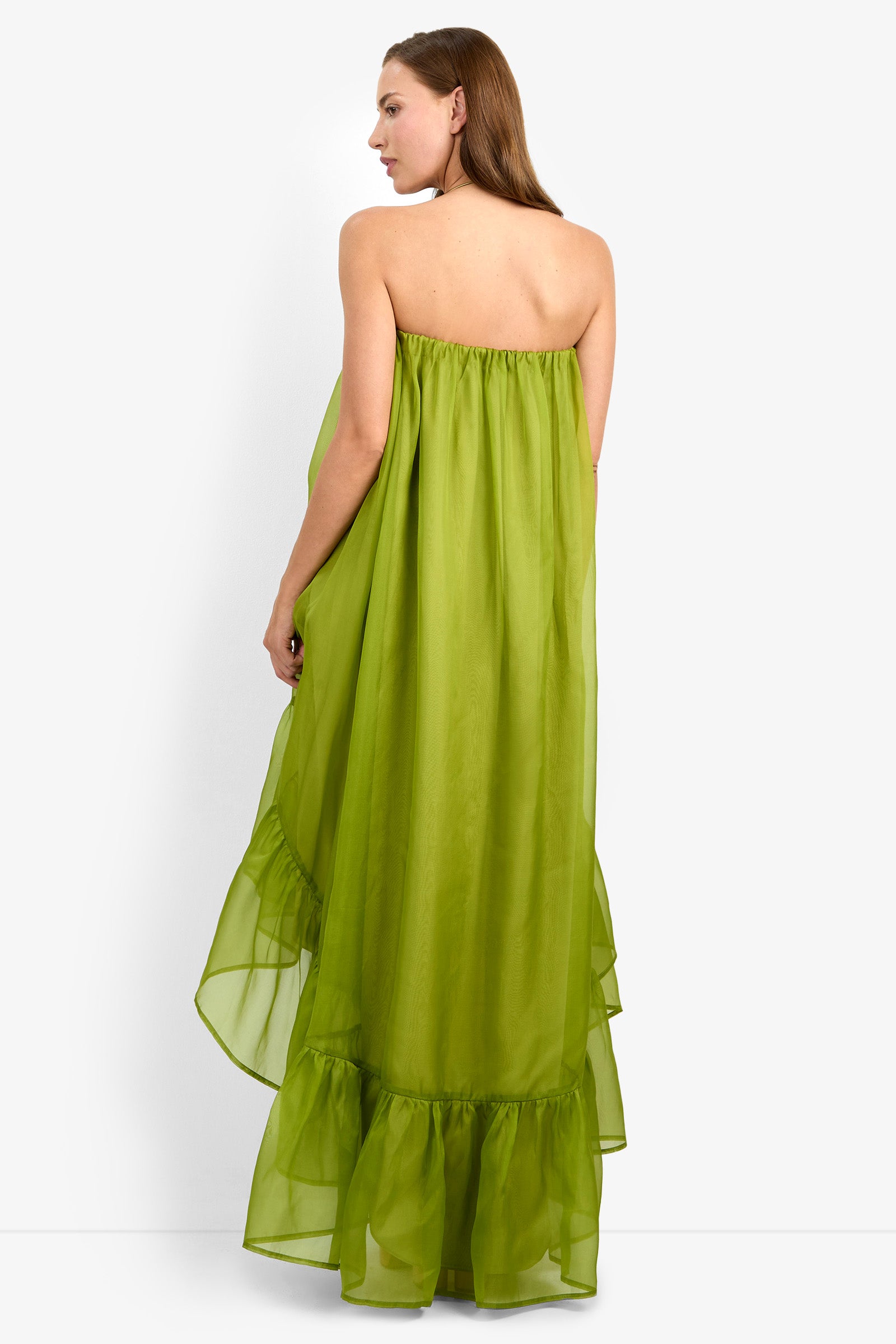 A woman with straight brown hair stands facing away against a plain white background, wearing the ARIAH DRESS—a strapless, long lime-green dress featuring a ruffled hem.