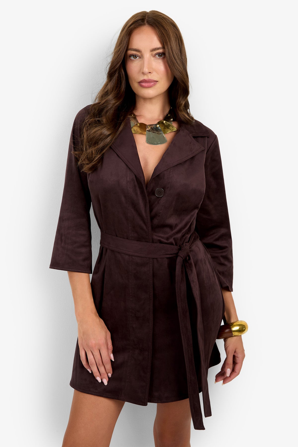 A woman stands against a plain white background wearing the Annecy Dress, a dark brown belted blazer dress with three-quarter sleeves, styled with a statement necklace and a large gold bracelet.