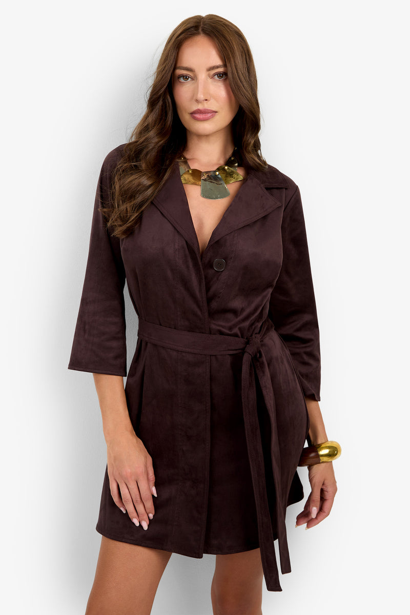 A woman stands against a plain white background wearing the Annecy Dress, a dark brown belted blazer dress with three-quarter sleeves, styled with a statement necklace and a large gold bracelet.