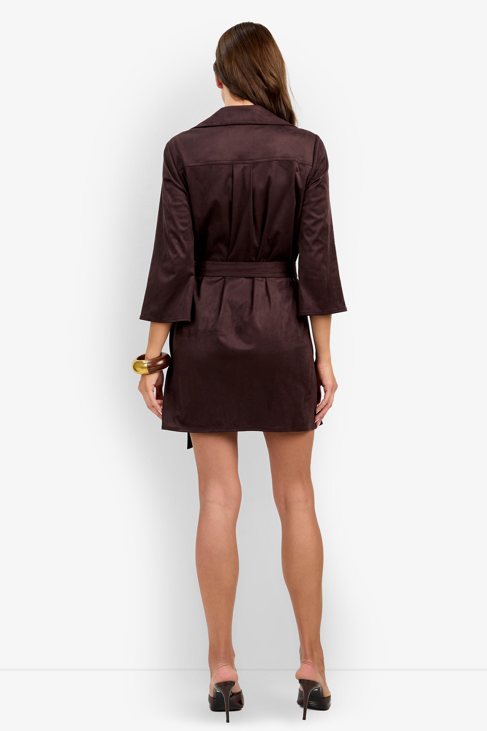 A woman stands facing away, wearing the Annecy Dress—a dark brown belted coat dress with three-quarter sleeves and black heeled shoes—holding a gold bracelet in her left hand.