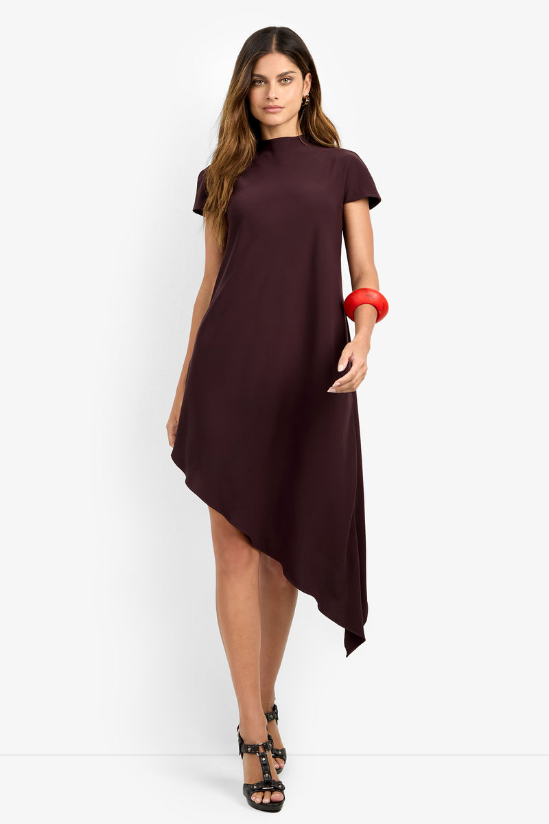 A woman stands against a plain white background wearing the SHANA 2 DRESS, an asymmetrical burgundy dress with short sleeves, paired with black high-heeled sandals and a large red bracelet on her right wrist.