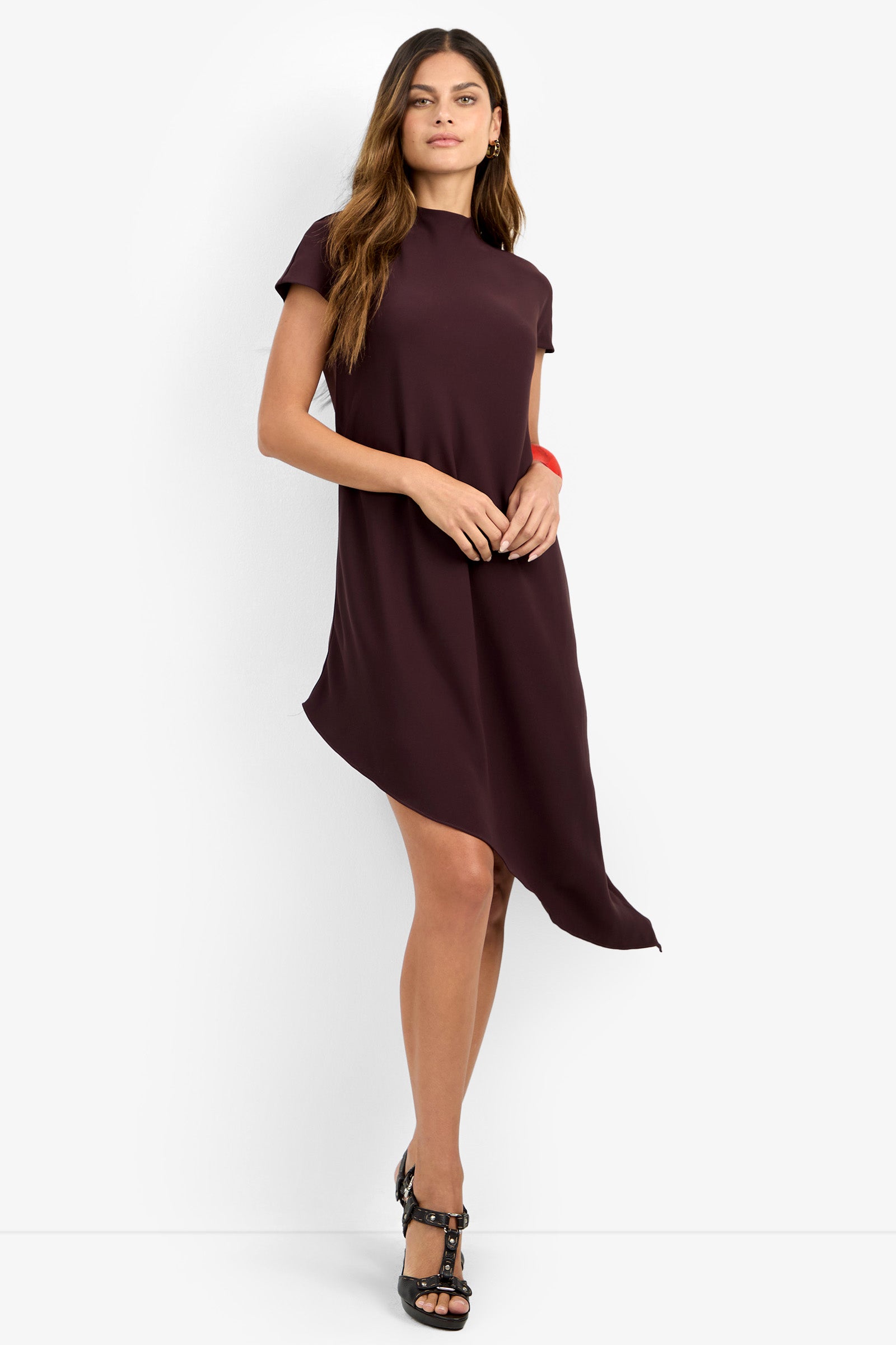 A woman stands confidently against a plain white background, wearing the SHANA 2 DRESS—a short-sleeve, dark maroon asymmetrical dress—paired with black heeled sandals and posing with one hand resting on her hip.