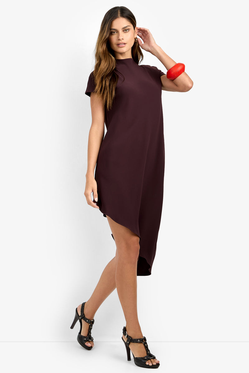 A woman wears the SHANA 2 DRESS, a short-sleeved, asymmetrical dark burgundy dress, paired with black high-heeled sandals and a bold red bracelet, as she walks confidently against a plain white background.