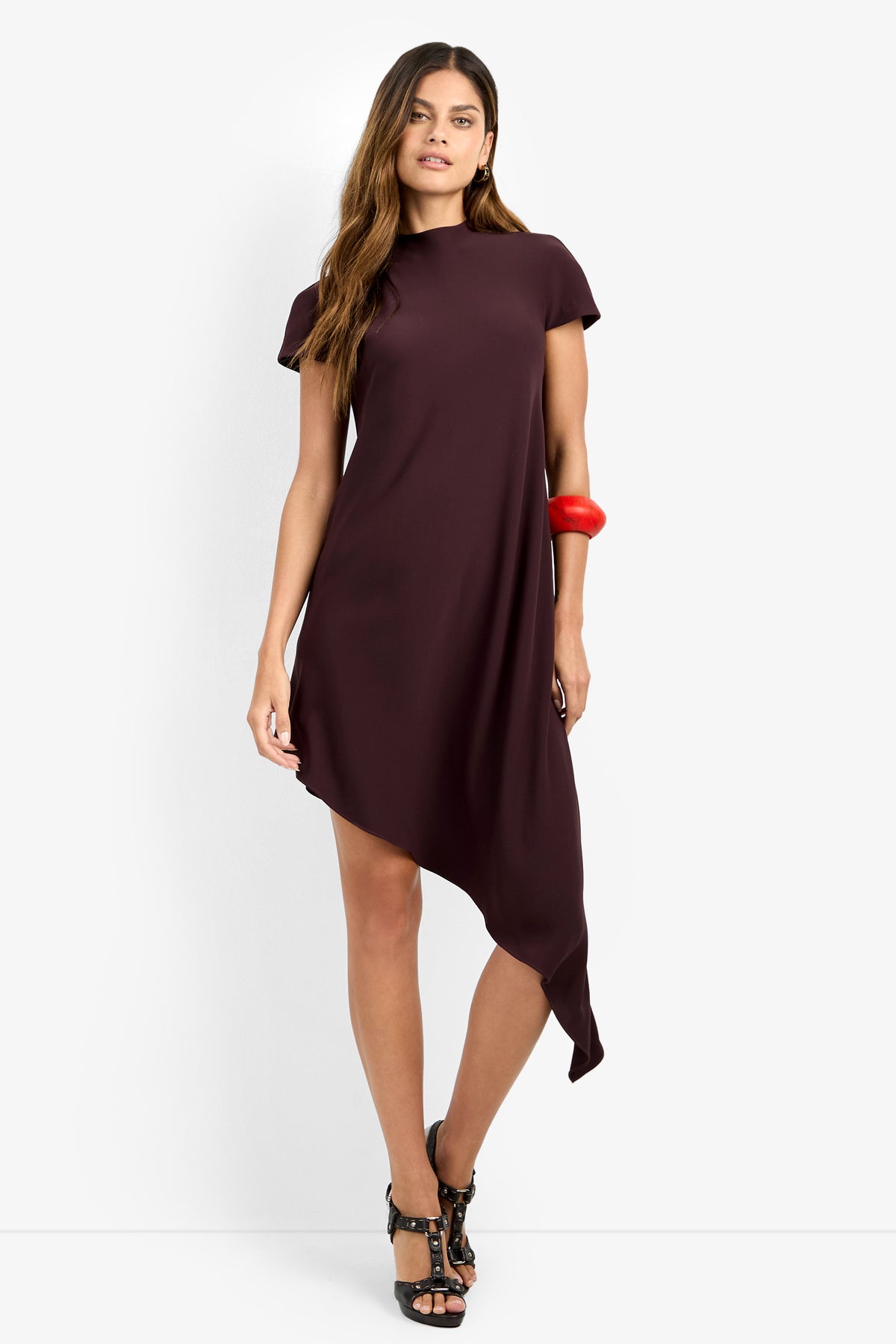 A woman stands against a plain white background wearing the SHANA 2 DRESS—a short-sleeved, dark maroon asymmetrical dress with an uneven hem—paired with black high-heeled sandals and a chunky red bracelet.