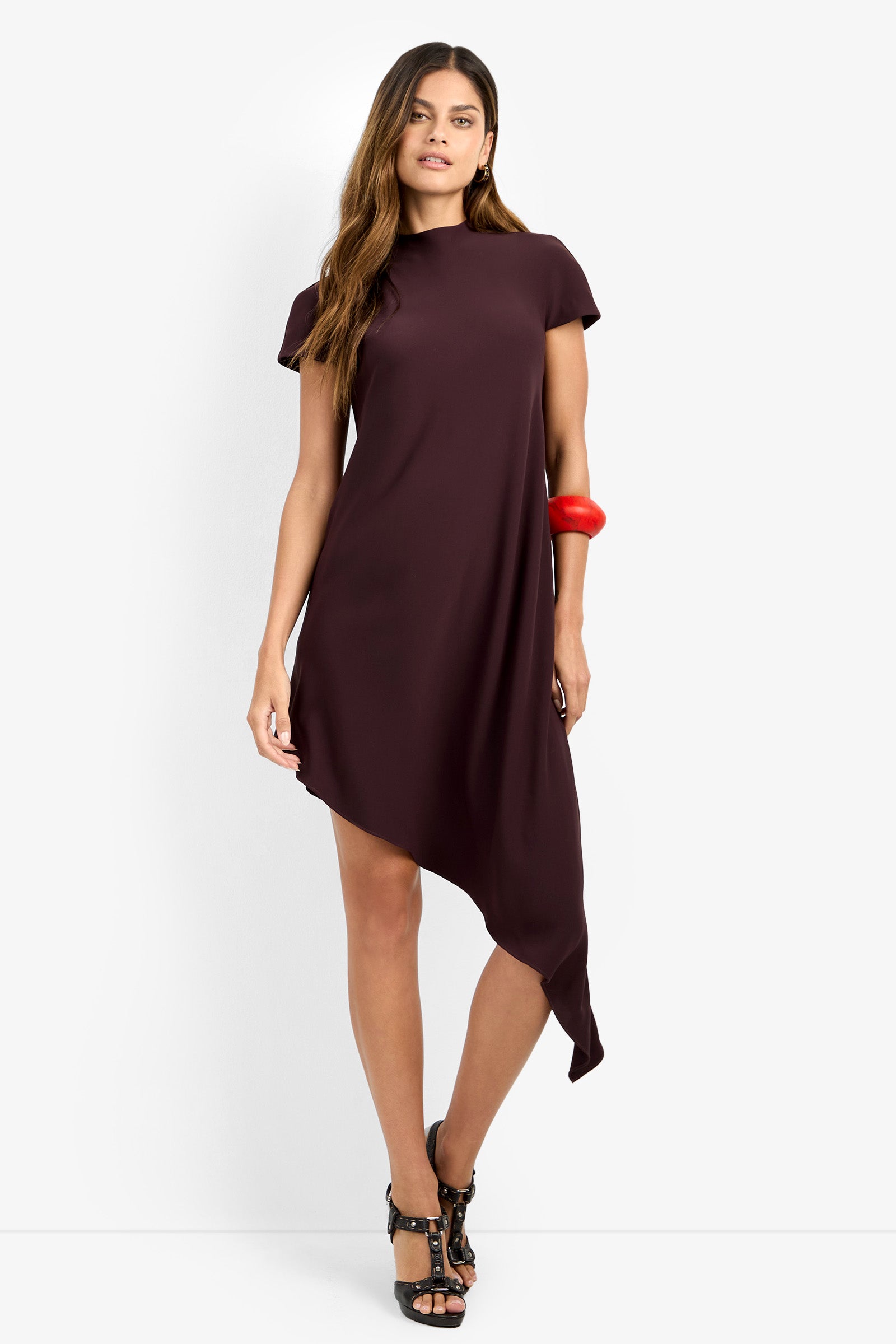A woman stands against a plain white background wearing the SHANA 2 DRESS—a short-sleeved, dark maroon asymmetrical dress with an uneven hem—paired with black high-heeled sandals and a chunky red bracelet.