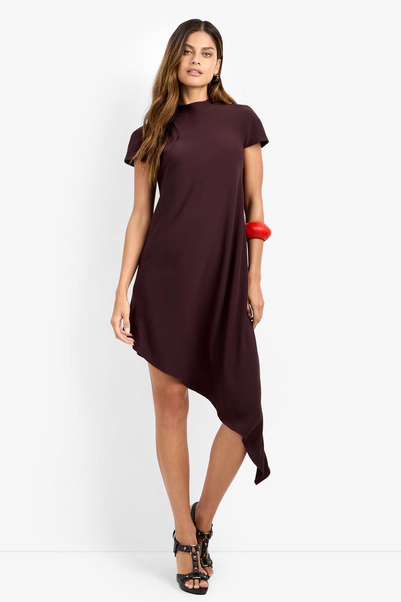 A woman stands against a plain white background wearing the SHANA 2 DRESS—a short-sleeved, dark maroon asymmetrical dress with an uneven hem—paired with black high-heeled sandals and a chunky red bracelet.