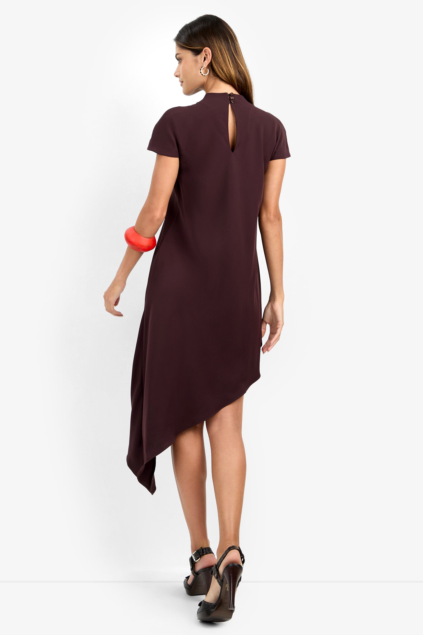 A woman wearing the SHANA 2 DRESS, a short-sleeve asymmetrical dark purple dress featuring a back keyhole detail, paired with black heels, hoop earrings, and a chunky red bracelet, stands against a white background.