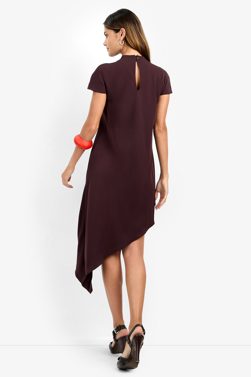 A woman wearing the SHANA 2 DRESS, a short-sleeve asymmetrical dark purple dress featuring a back keyhole detail, paired with black heels, hoop earrings, and a chunky red bracelet, stands against a white background.