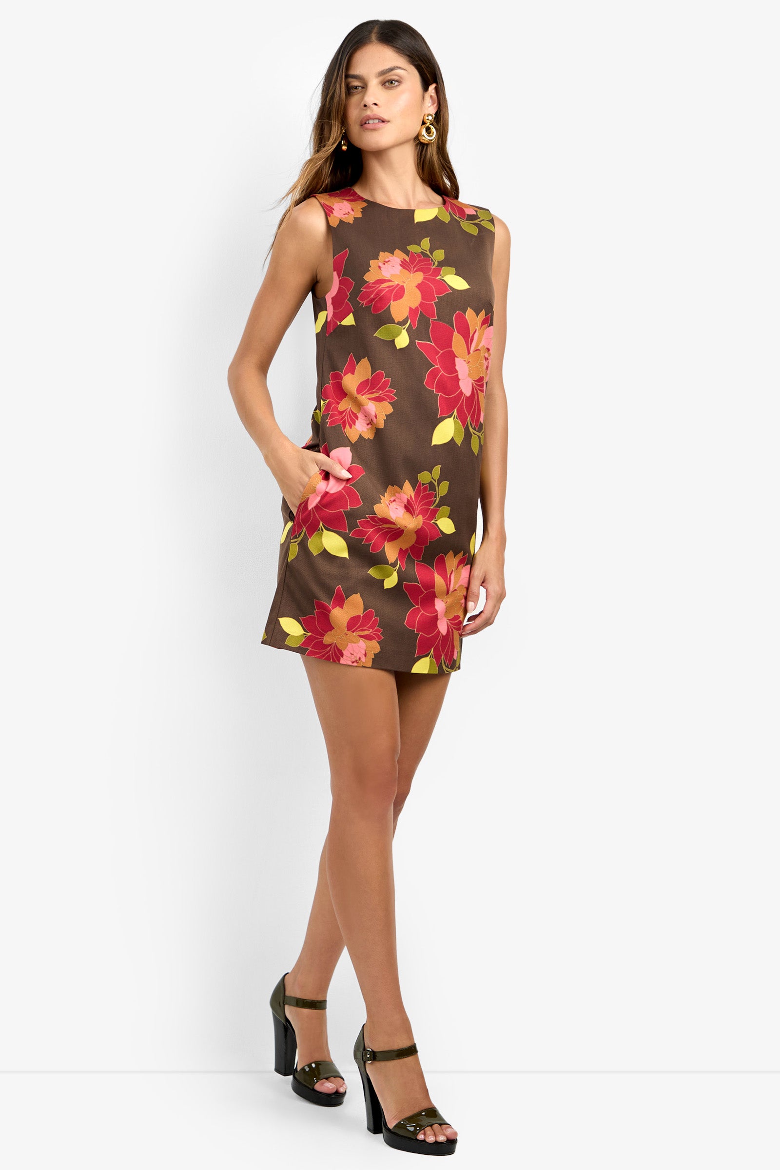 A woman models the TAHLIA 2 DRESS, a sleeveless brown mini dress featuring bold red and pink floral prints. She pairs it with green earrings and dark green high-heeled sandals, standing against a white background while gazing slightly to the side.