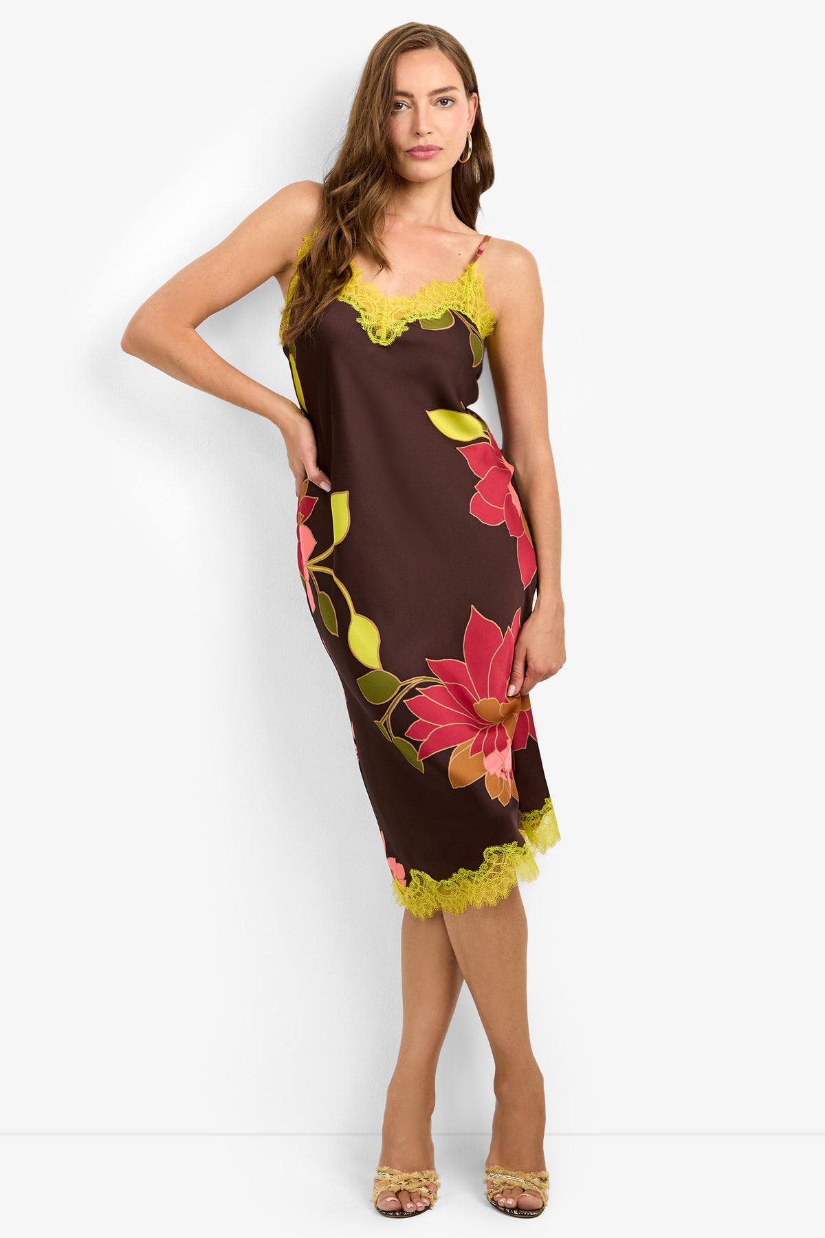 Model wearing the CARAMIA DRESS (1), a brown slip dress featuring large pink and yellow floral prints and yellow lace trim, standing with one hand on her hip and the other by her side against a plain white background.