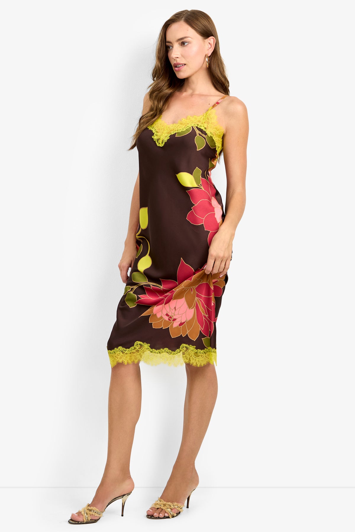 A woman wears the CARAMIA DRESS (1), a sleeveless brown slip dress featuring yellow lace trim and a bold pink floral print, as she stands in heels against a plain white background.