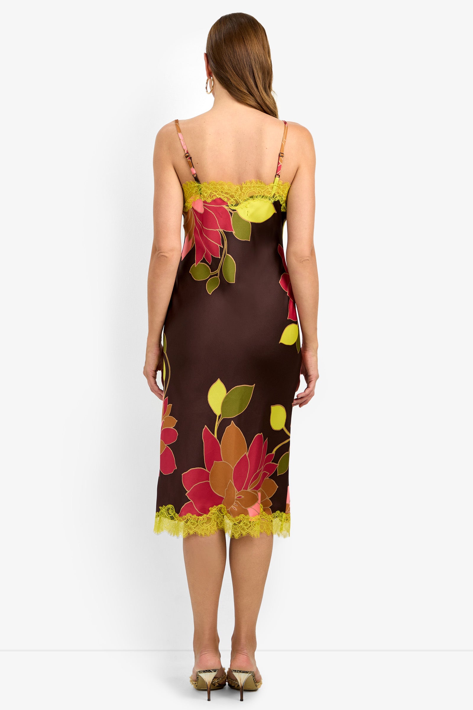 A woman with straight, long brown hair is seen from the back wearing the CARAMIA DRESS (1), a brown midi slip dress featuring bright red, yellow, and green floral patterns, yellow lace trim, and thin straps. She pairs the look with beige heeled sandals.