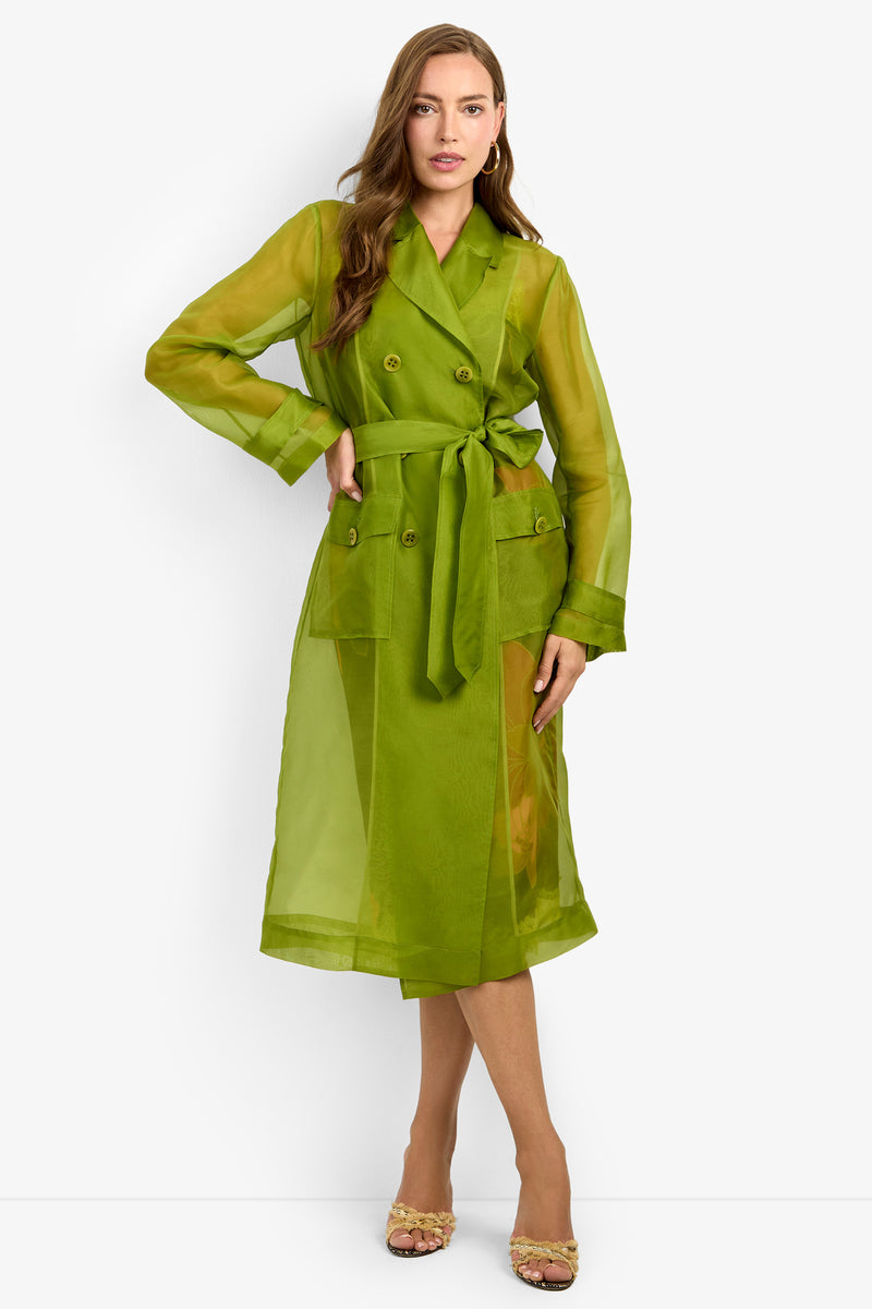 A woman poses in the ANGELICO TRENCH, a sheer, bright green, double-breasted coat with a matching belt, styled with gold heeled sandals against a plain white background.