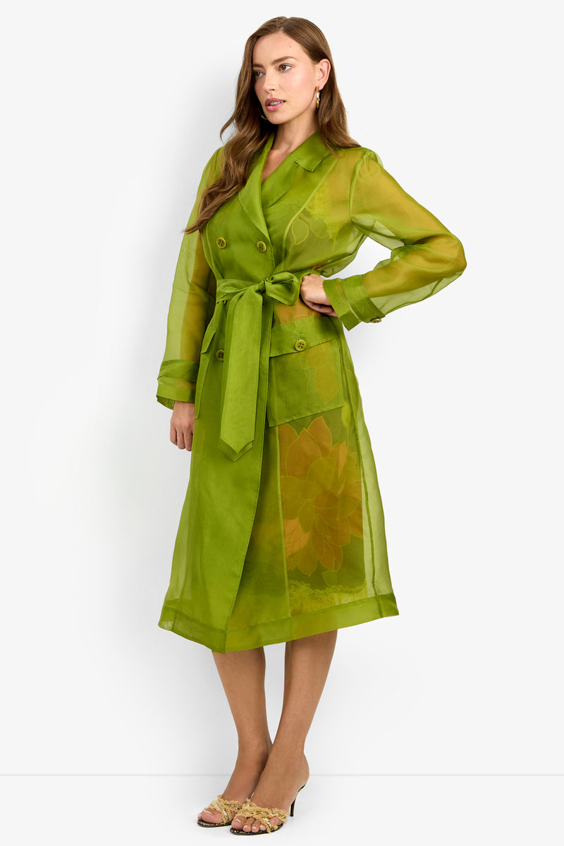 Wearing the ANGELICO TRENCH—a sheer, green trench coat with long sleeves, a belt at the waist, and floral details—paired with open-toe heels, a woman stands against a plain white background.