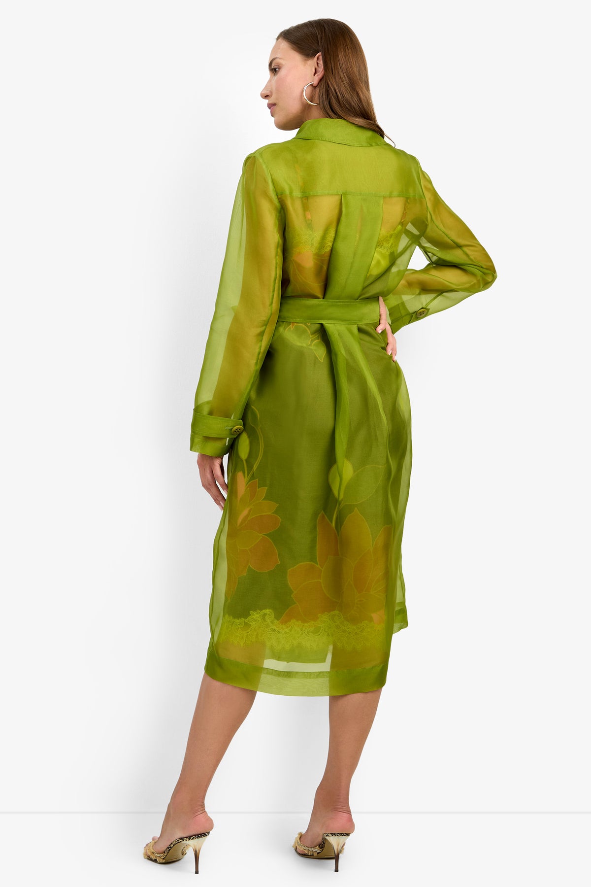 A woman stands with her back to the camera wearing the ANGELICO TRENCH, a sheer green belted dress with yellow floral patterns and long sleeves, paired with heeled sandals, against a plain white background.