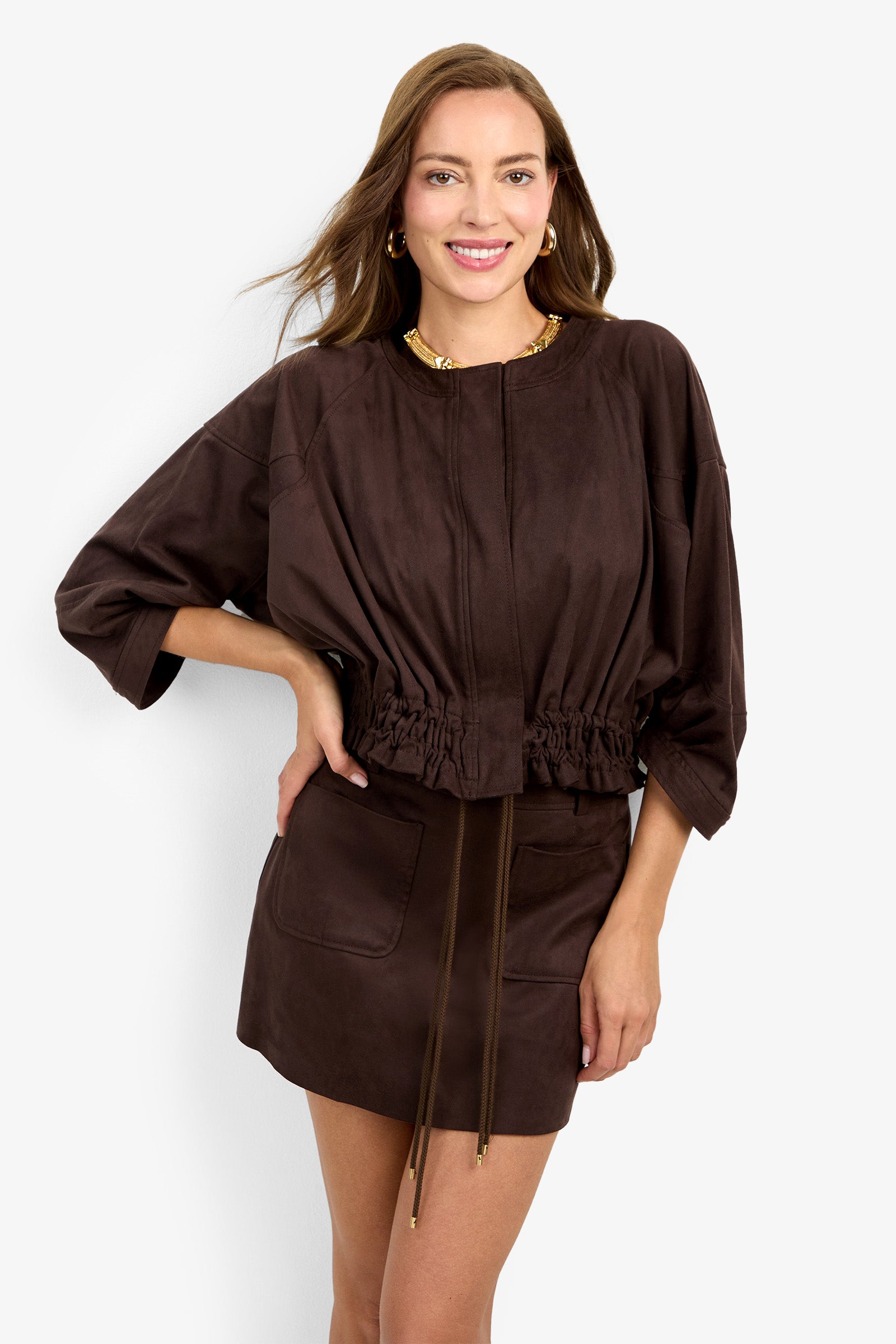 A woman is smiling and standing against a white background, wearing the PRITA JACKET in dark brown with loose sleeves and a drawstring waist, paired with a matching brown mini skirt.
