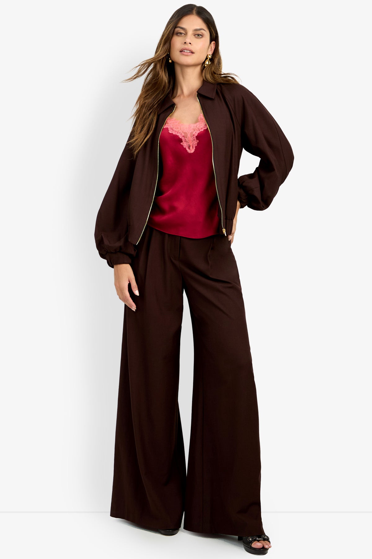 A woman stands with one hand on her hip against a plain white background, wearing the INFLUENTIAL JACKET—a loose brown jacket paired with matching pants over a red satin camisole.