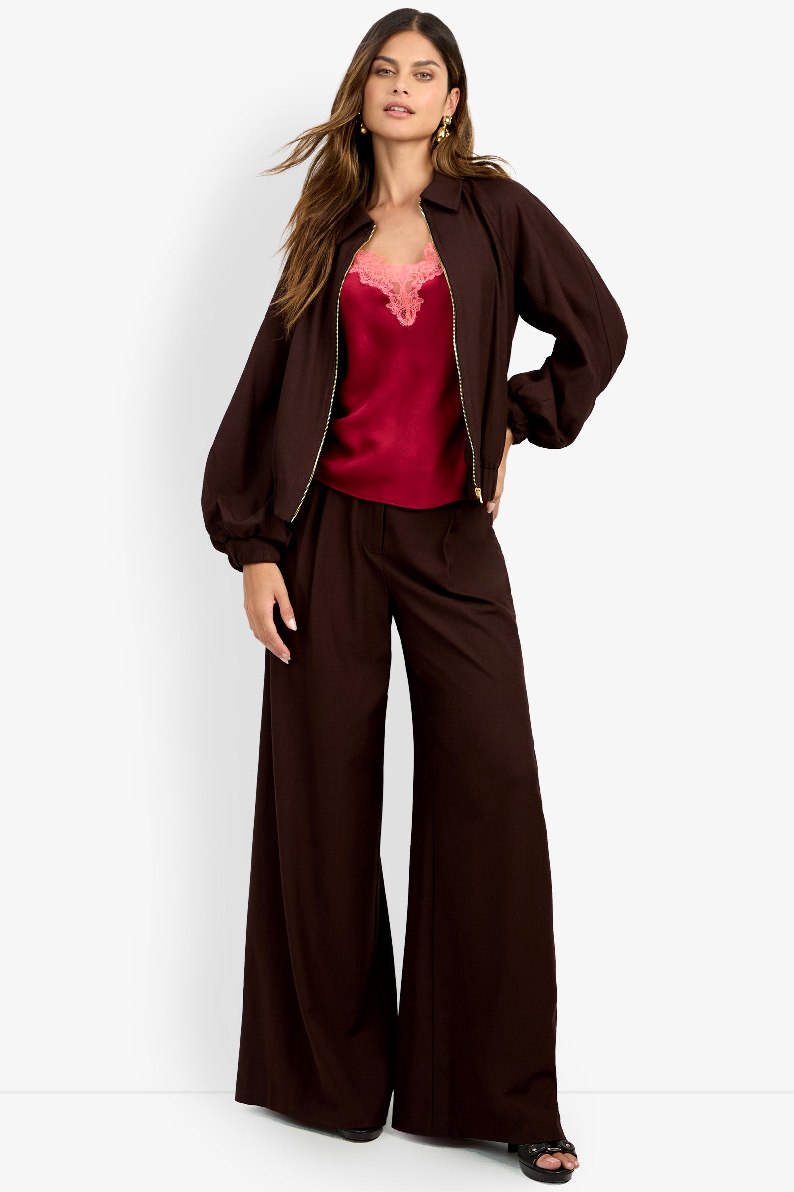 A woman stands with one hand on her hip against a plain white background, wearing the INFLUENTIAL JACKET—a loose brown jacket paired with matching pants over a red satin camisole.