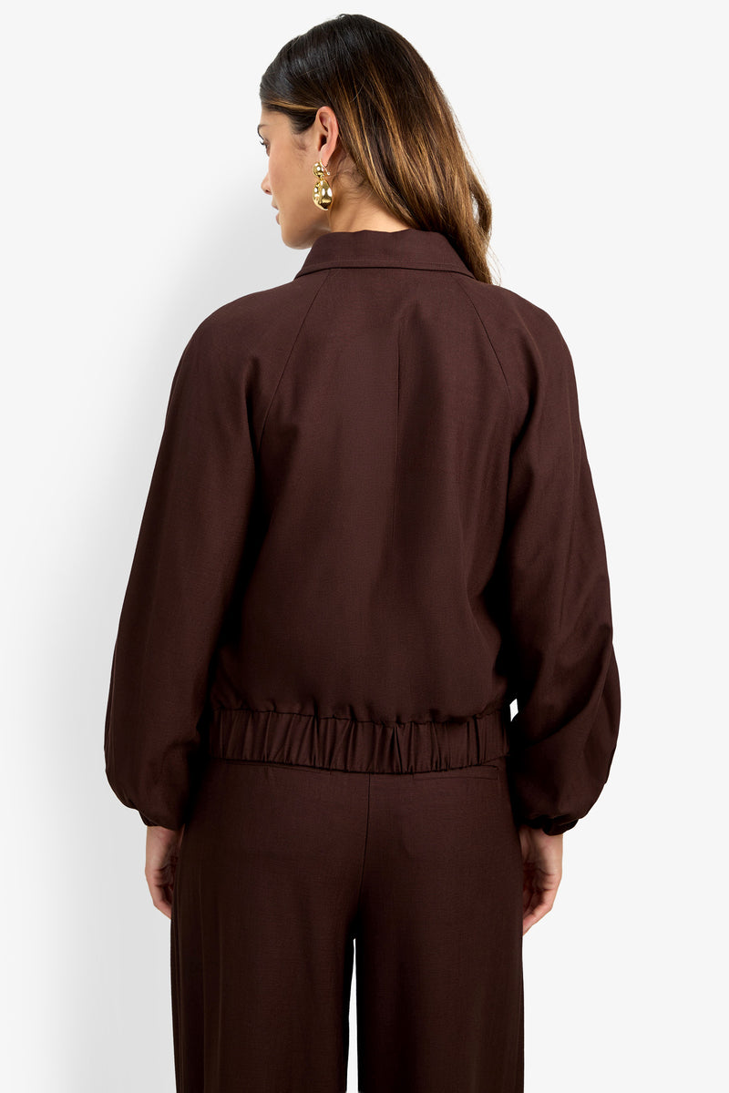 A woman with long brown hair is seen from the back wearing the INFLUENTIAL JACKET—a dark brown, loose-fitting piece with a gathered waist—paired with matching pants and gold earrings.