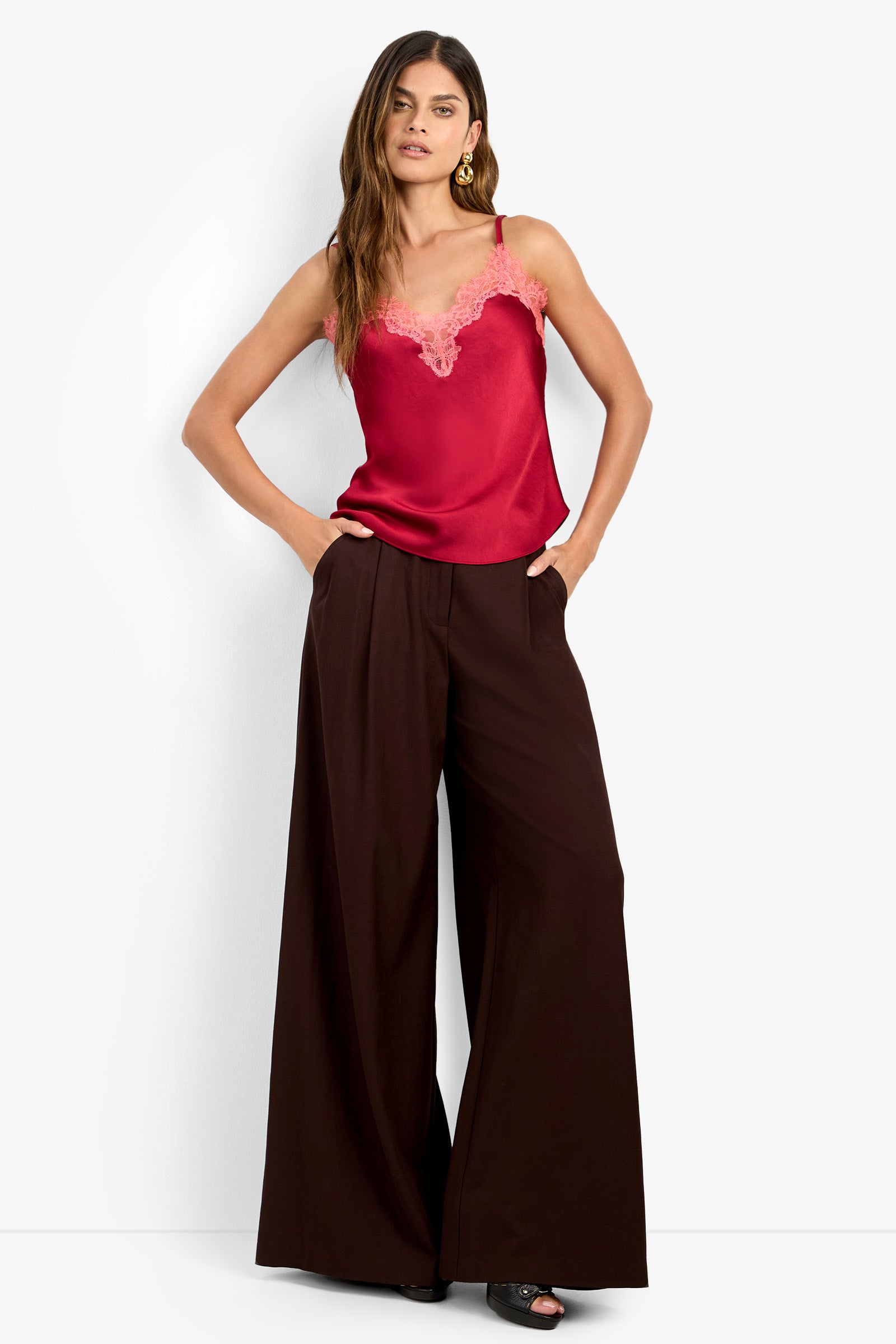 Woman with long hair wears a red satin lace-trim camisole and the MIMMO PANT, high-waisted, wide-leg brown trousers. She accessorizes with gold hoop earrings and black open-toe shoes, standing with hands in pockets.