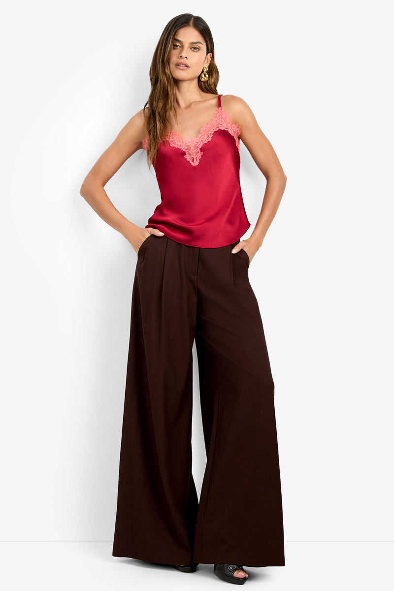 Woman with long hair wears a red satin lace-trim camisole and the MIMMO PANT, high-waisted, wide-leg brown trousers. She accessorizes with gold hoop earrings and black open-toe shoes, standing with hands in pockets.