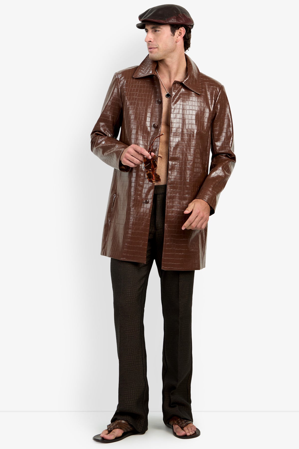 A man stands holding sunglasses, his shirt open to reveal his chest, wearing the Morton Trench Coat in brown faux-crocodile, paired with dark pants, sandals, and a flat cap.