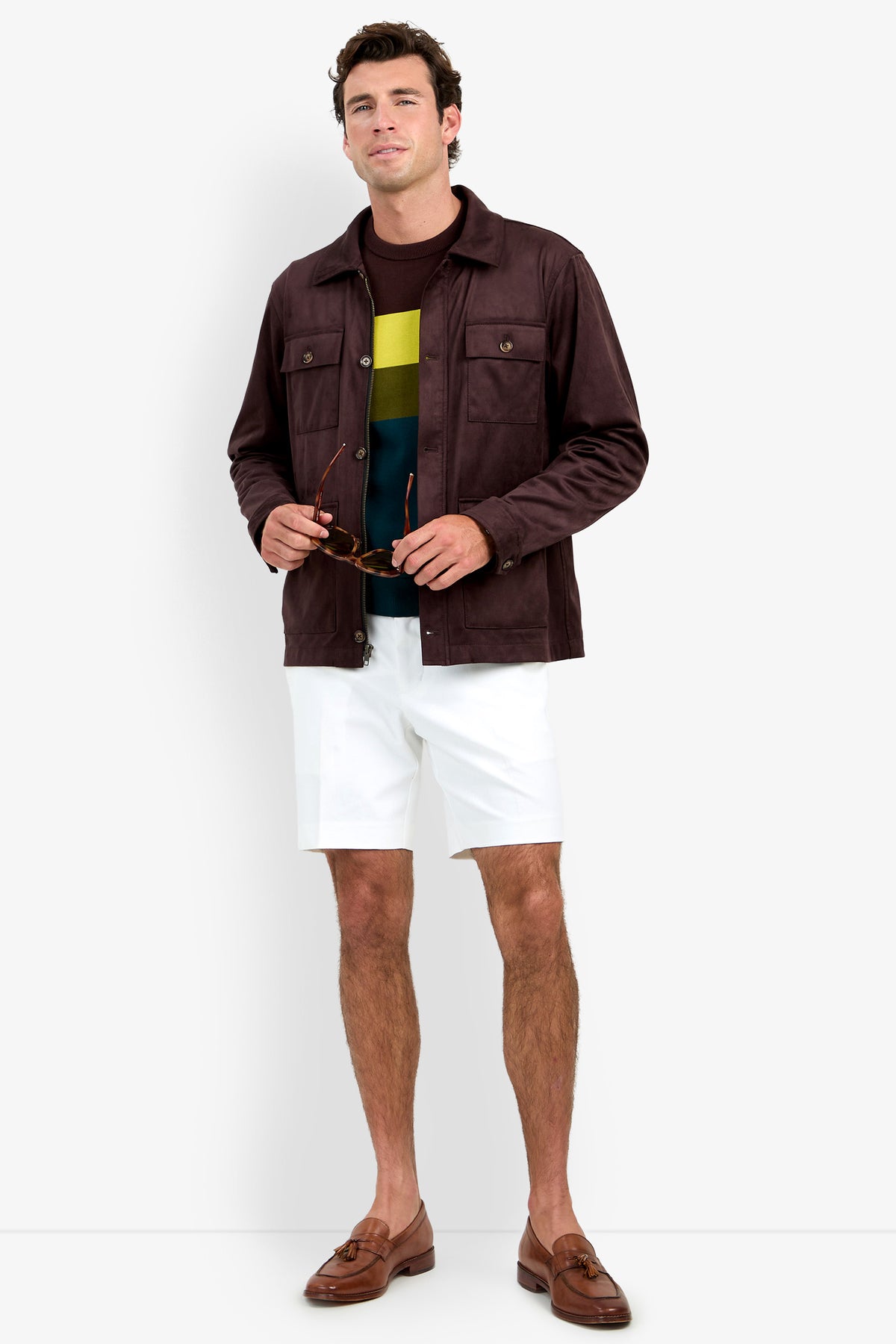 A man wearing the Highland Park Jacket, a multicolored striped shirt, white shorts, and brown loafers stands holding sunglasses and faces forward against a plain background.