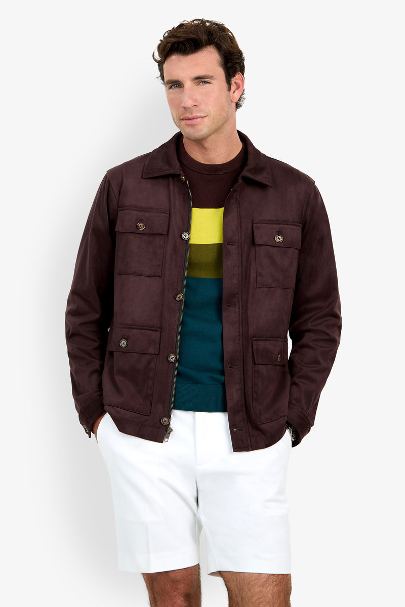 A man stands against a plain white background, smiling slightly with his hands in his pockets, wearing the Highland Park Jacket over a striped sweater and white shorts.