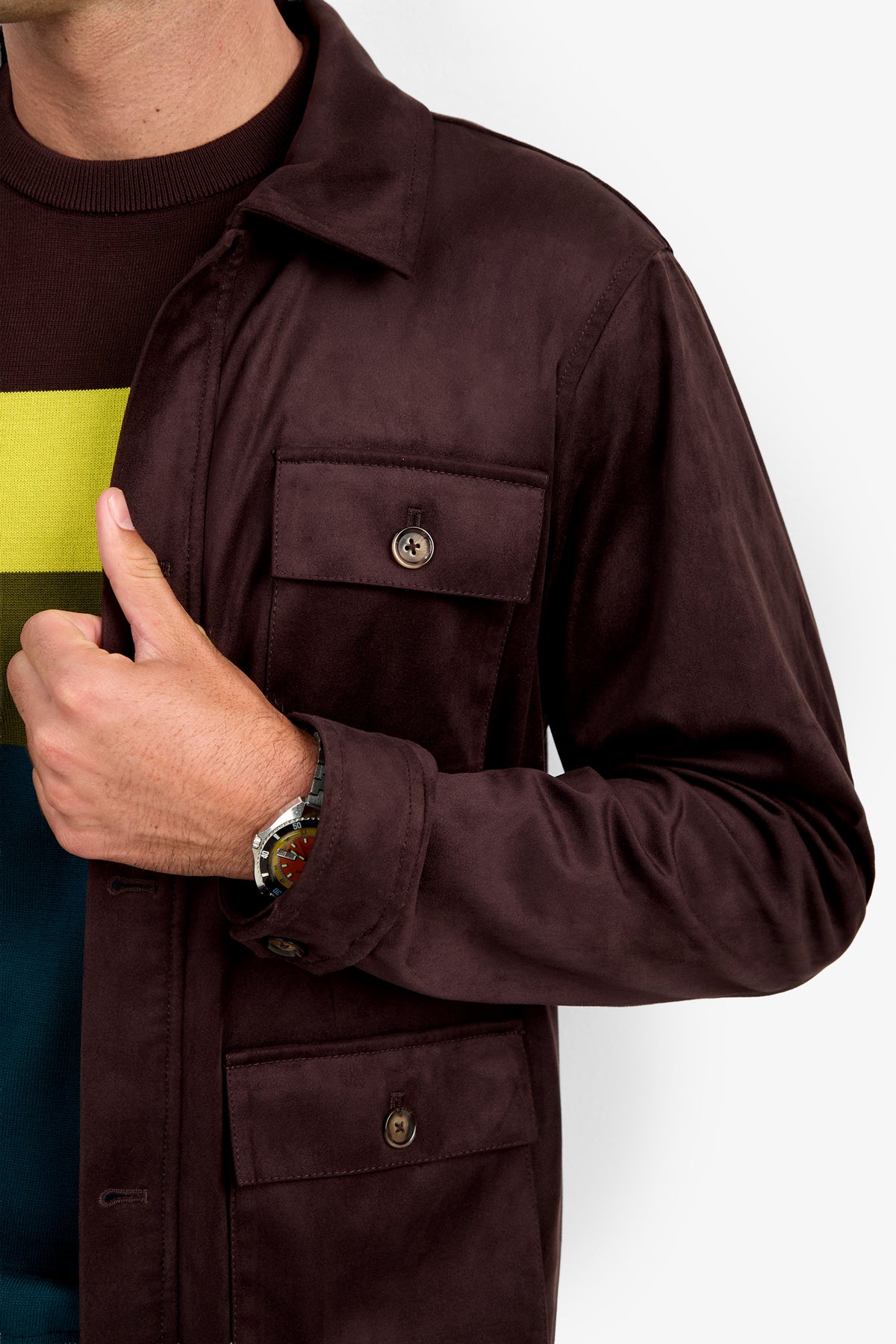 A person wearing the Highland Park Jacket over a colorful sweater, adjusting the jacket lapel with their left hand. A silver wristwatch is visible on their wrist against a plain white background.