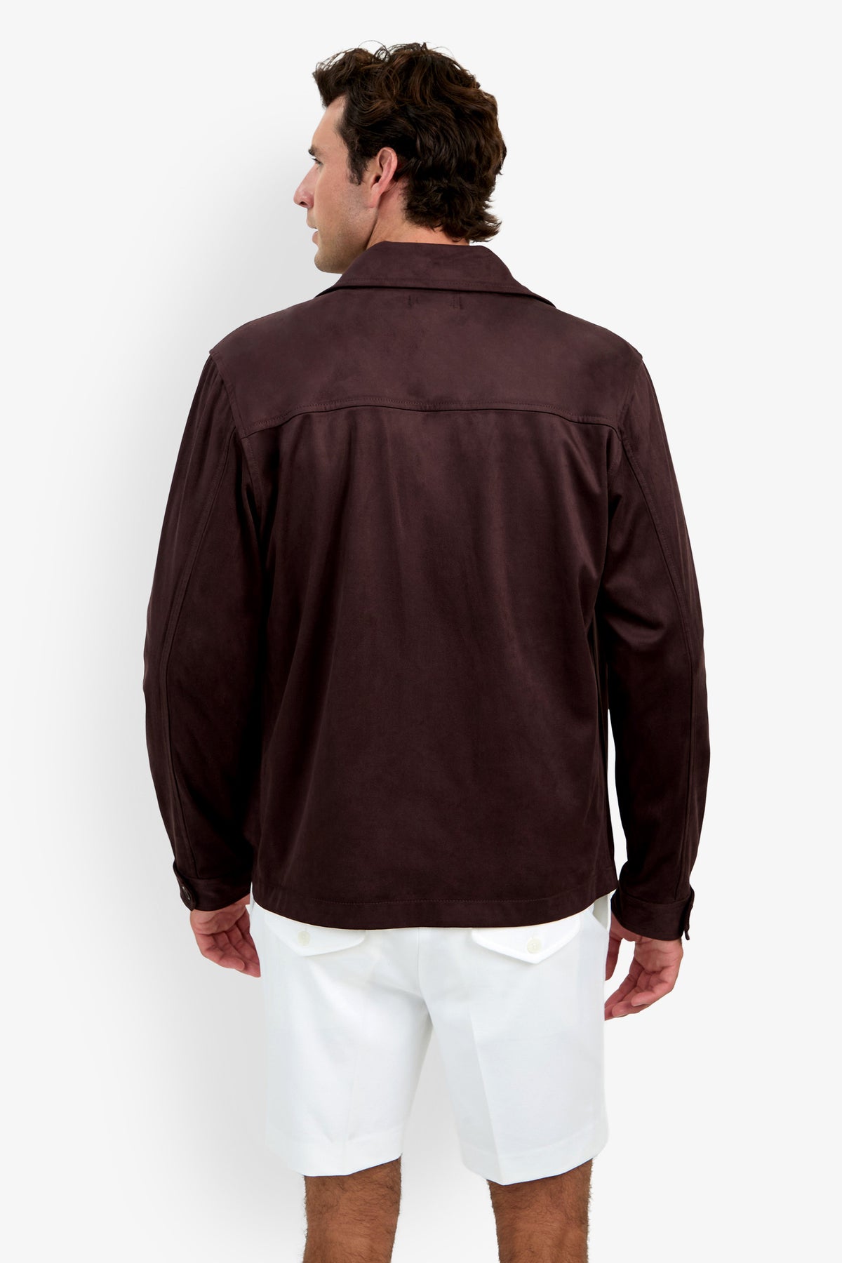 A man is standing with his back to the camera, wearing the Highland Park Jacket in dark brown along with white shorts against a plain light background.