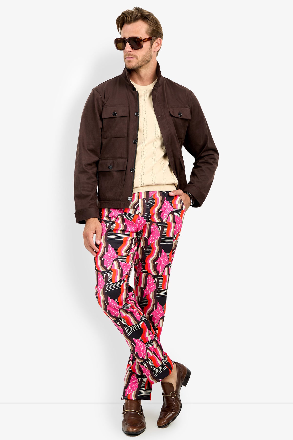 A man wearing sunglasses, the HIGHLAND PARK JACKET, a cream sweater, vibrant pink patterned pants, and brown shoes stands against a plain light background.