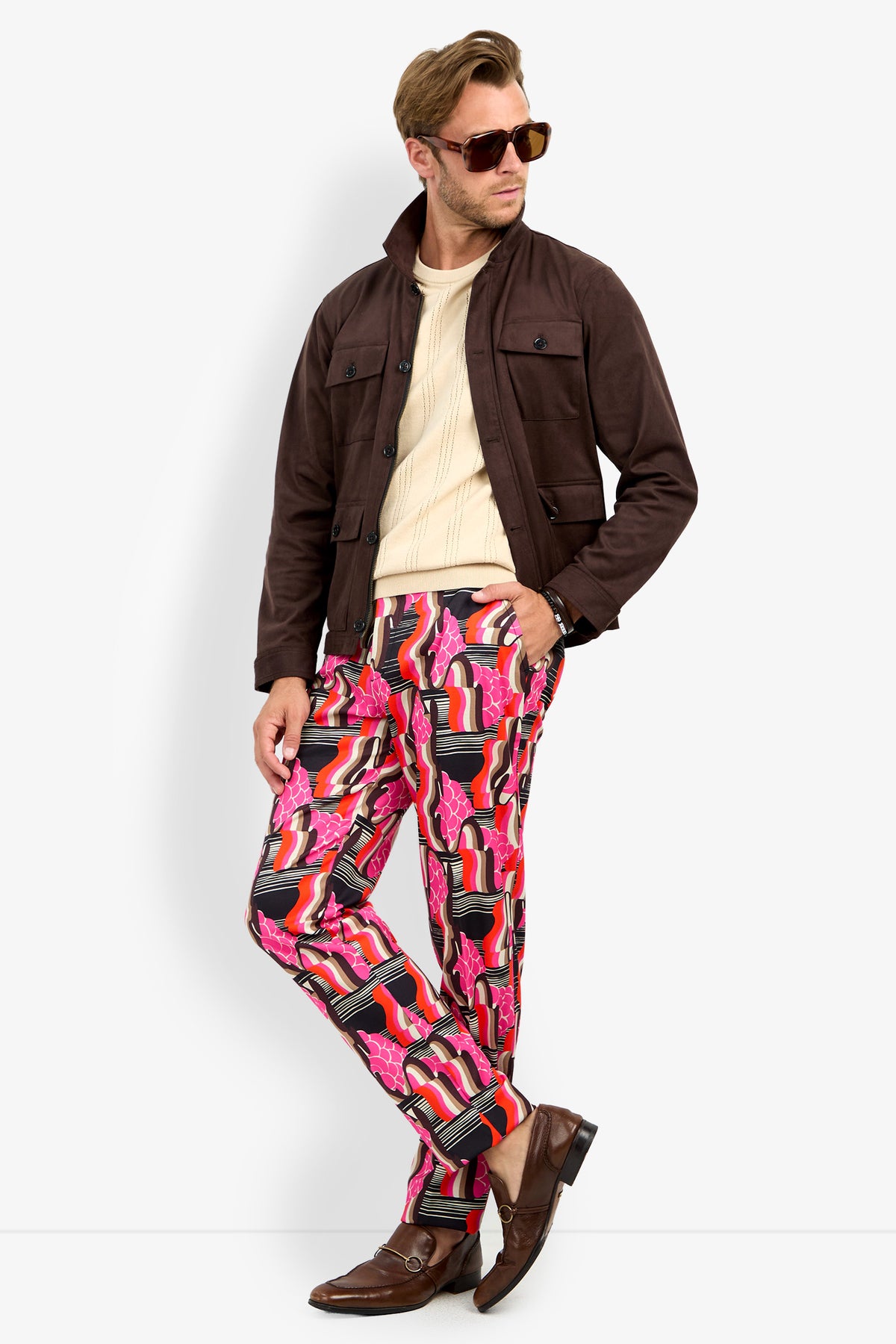A man wearing sunglasses, the HIGHLAND PARK JACKET in brown, a cream sweater, and colorful patterned pants featuring red, pink, and black designs stands confidently with crossed legs and brown loafers.