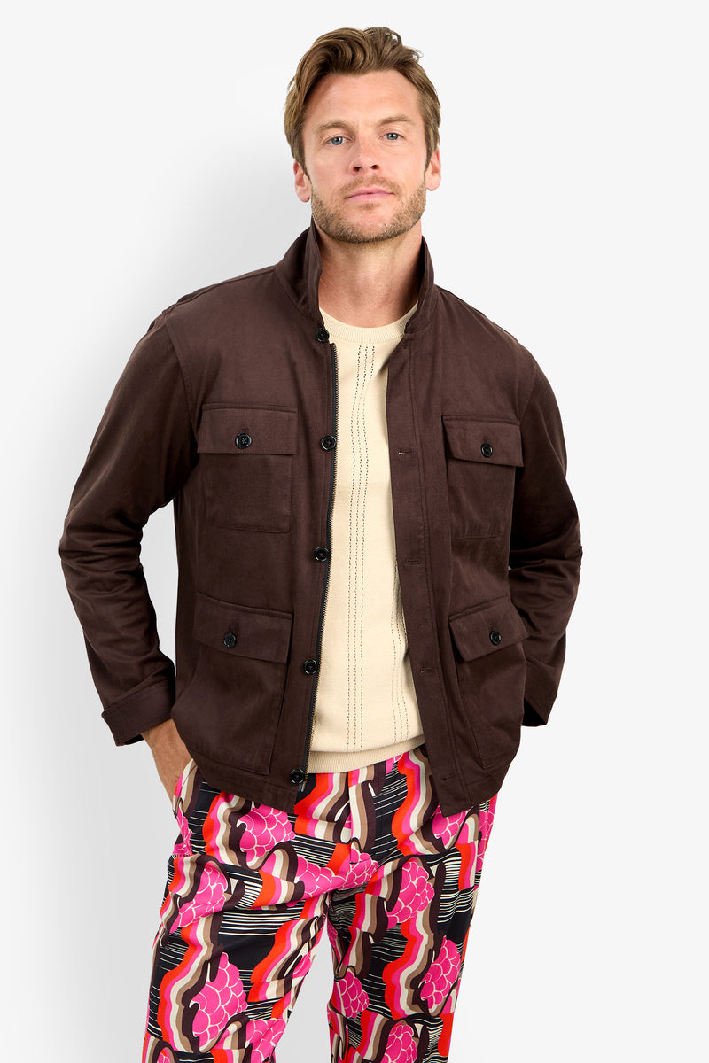 A man wearing the HIGHLAND PARK JACKET over a cream sweater and bright, patterned pink and black pants stands with his hands in his pockets against a plain light background.
