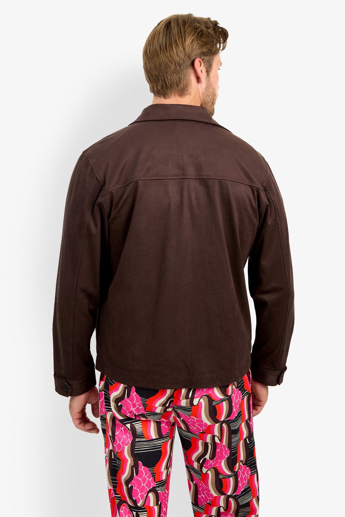 A man stands facing away, wearing the HIGHLAND PARK JACKET—a dark brown jacket—paired with colorful, patterned pants featuring pink, red, black, and white swirls and shapes, set against a plain white background.