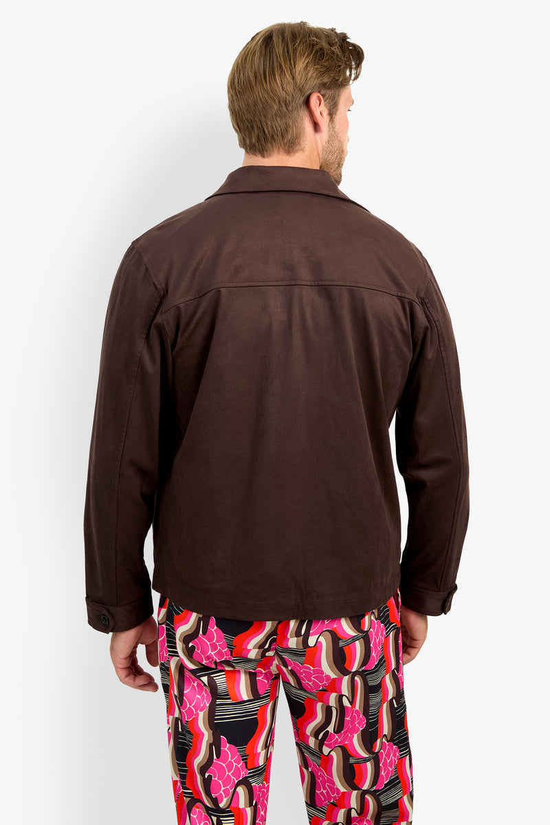 A man stands facing away, wearing the HIGHLAND PARK JACKET—a dark brown jacket—paired with colorful, patterned pants featuring pink, red, black, and white swirls and shapes, set against a plain white background.