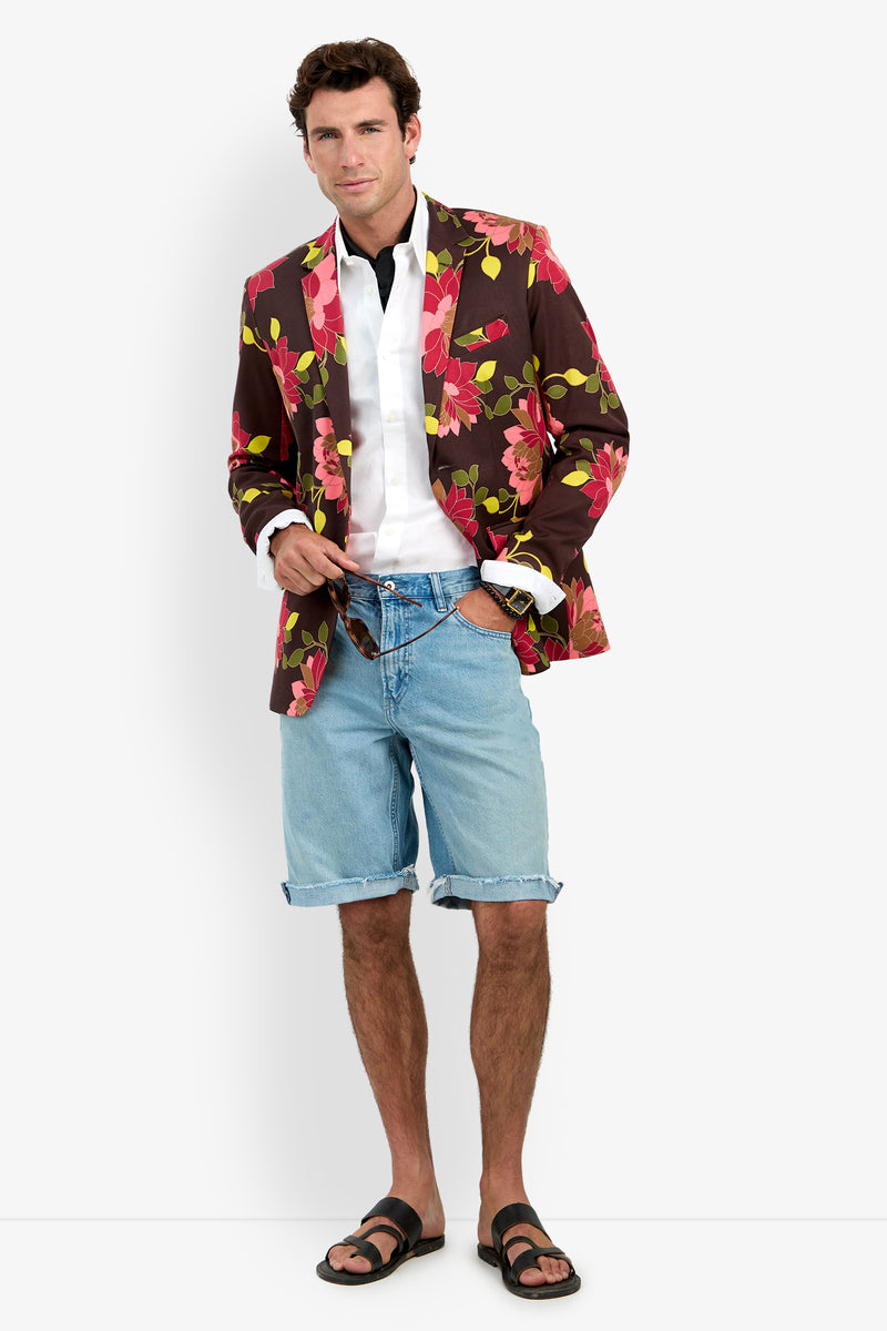 A man stands against a plain background in a relaxed pose and a slight smile, wearing the Thurston Blazer, paired with a white shirt, blue denim shorts, and sandals.