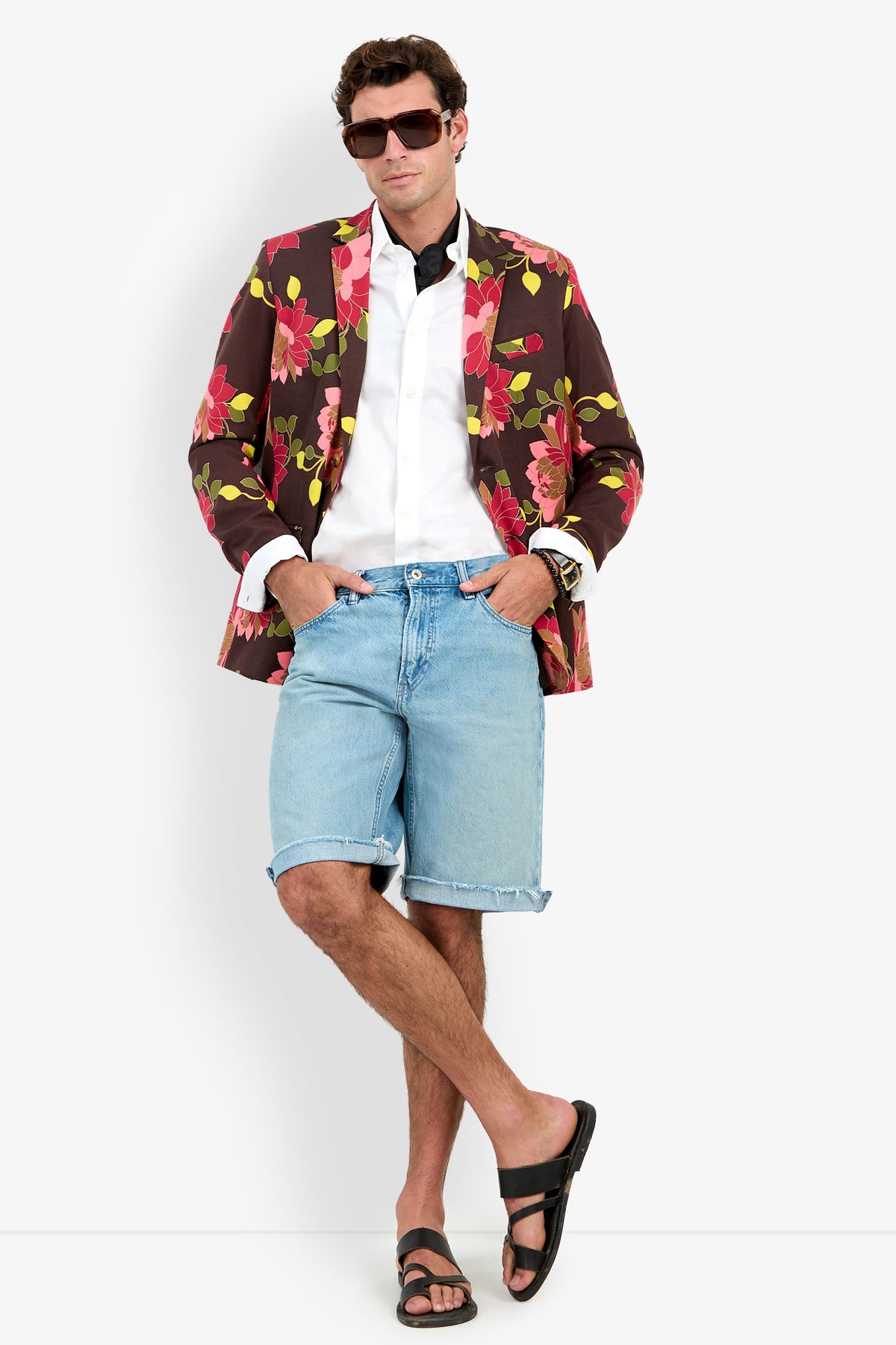 A man wearing sunglasses, the Thurston Blazer over a white shirt, denim shorts, and black sandals stands with legs crossed and hands in pockets against a plain background.