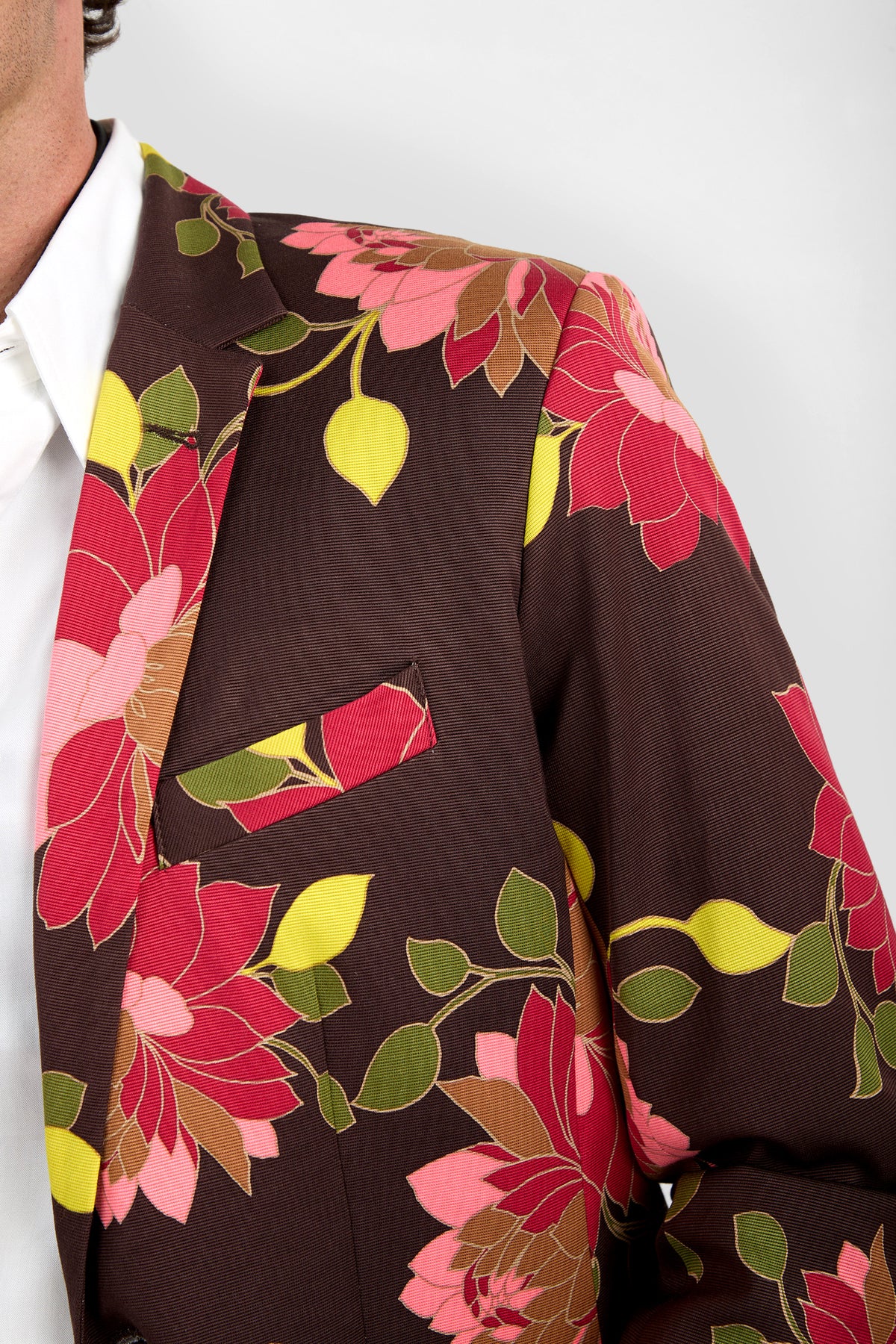 A close-up of a person wearing the Thurston Blazer, which features a brown base with a vibrant floral pattern in red, pink, and yellow. The shirt underneath is white. Only the right side of the blazer and part of the face are visible.