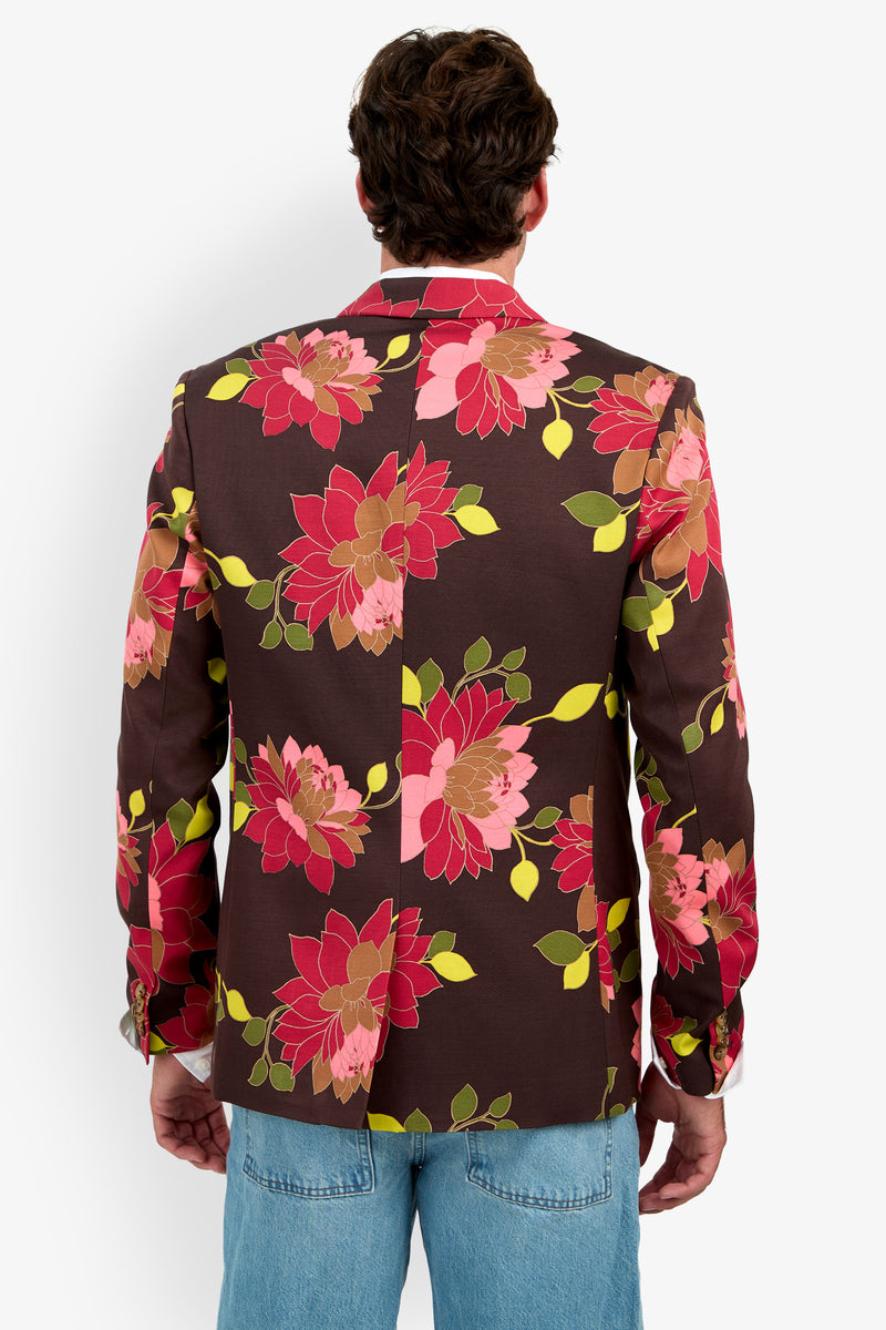A person wearing the Thurston Blazer, featuring large red, pink, and green floral patterns on a brown background, paired with light blue jeans and shown from the back against a plain white background.