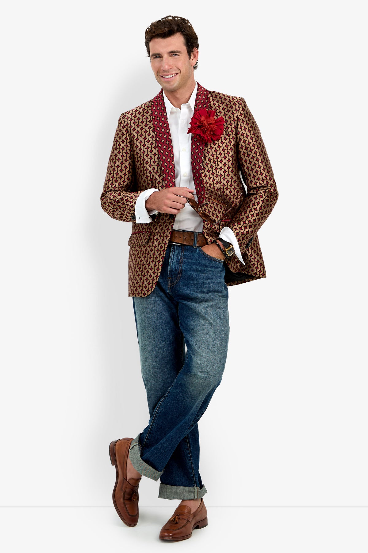A man with short brown hair stands smiling, wearing a white button-down shirt, cuffed blue jeans, and brown leather loafers. He is styled in the Alfred Blazer—a patterned gold and red jacket—adorned with a red flower lapel pin, and holds sunglasses in one hand. White background.