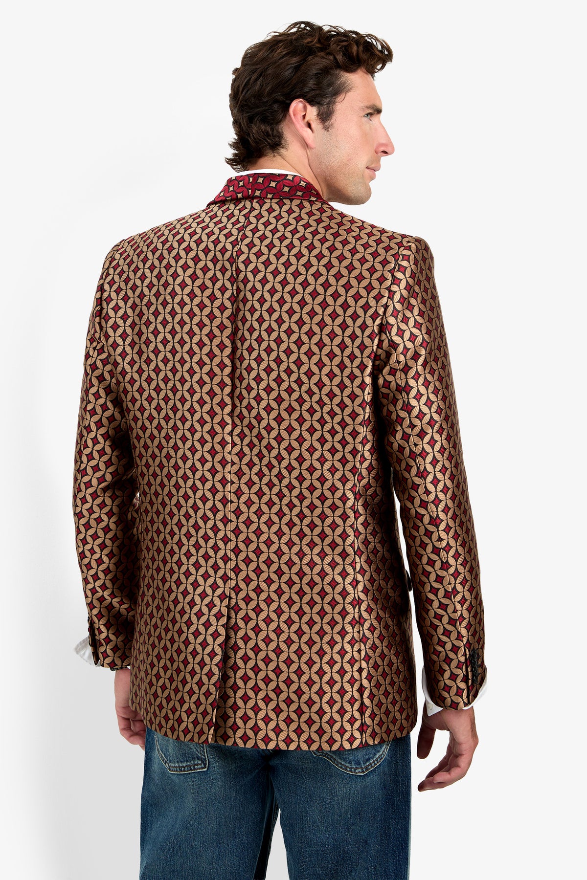 A man wearing the Alfred Blazer—a patterned gold and red blazer—paired with blue jeans stands facing away, looking to the side against a plain white background.