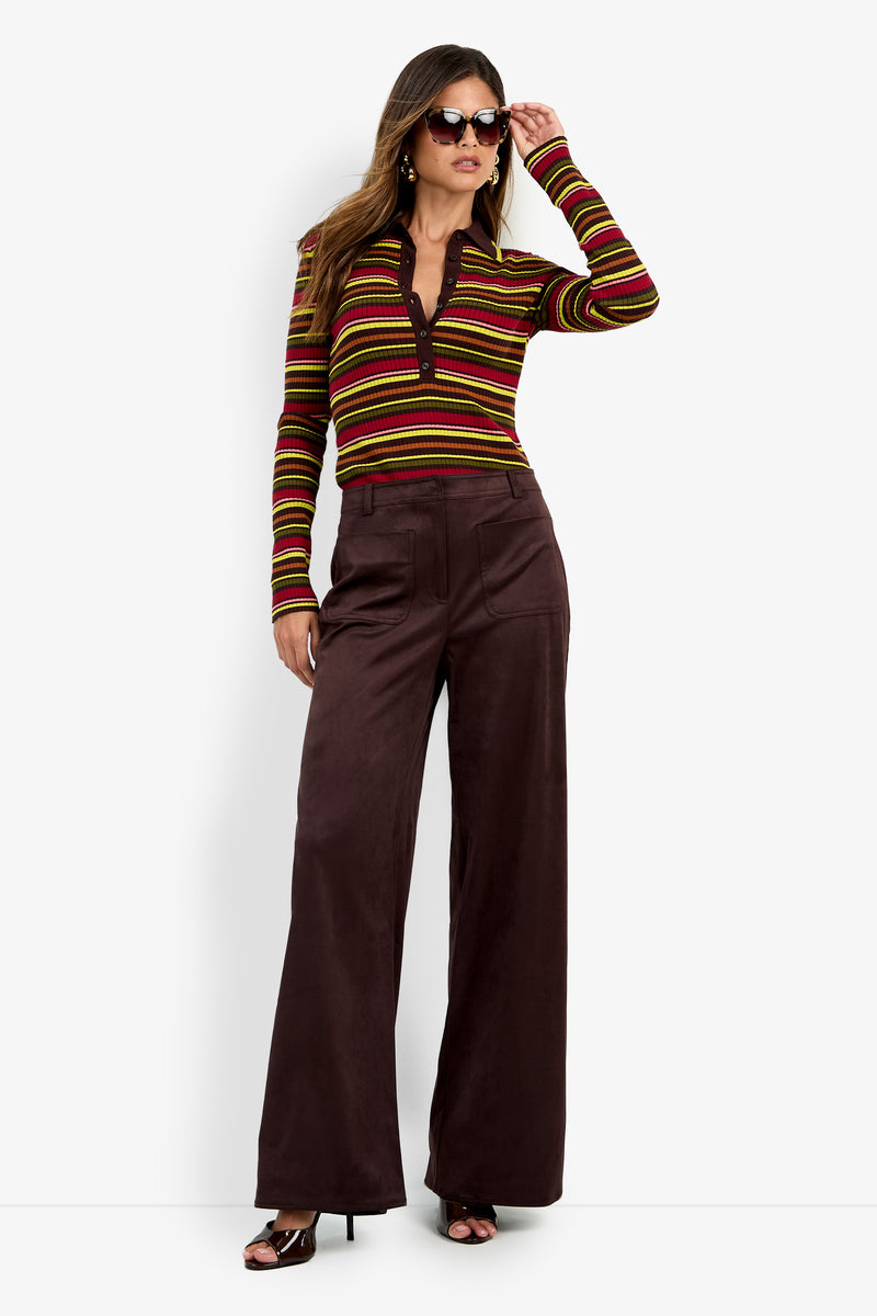 Woman wearing sunglasses, a long-sleeve striped shirt with red, yellow, and green tones, and the Zinn Pant—wide-leg dark brown pants—standing and posing with one hand on her hip.