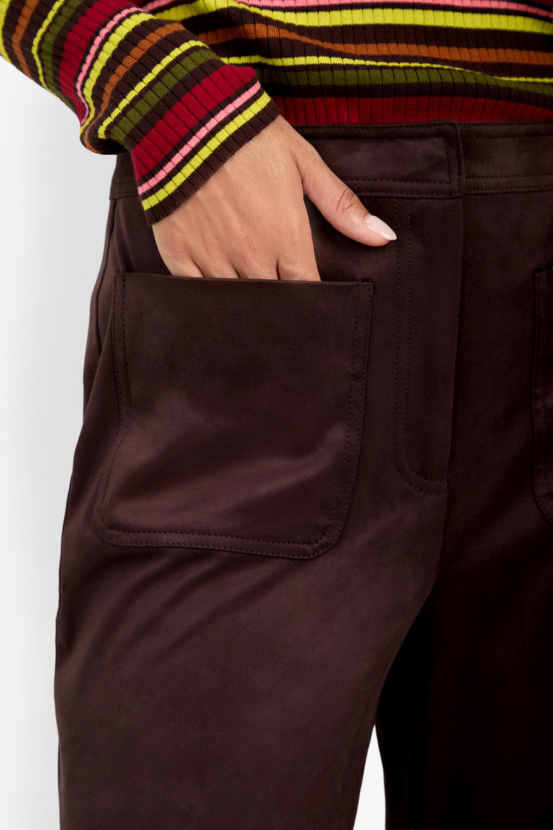 A person wearing a colorful striped sweater and the Zinn Pant in dark brown, featuring large front pockets, with their hand resting inside one pocket.