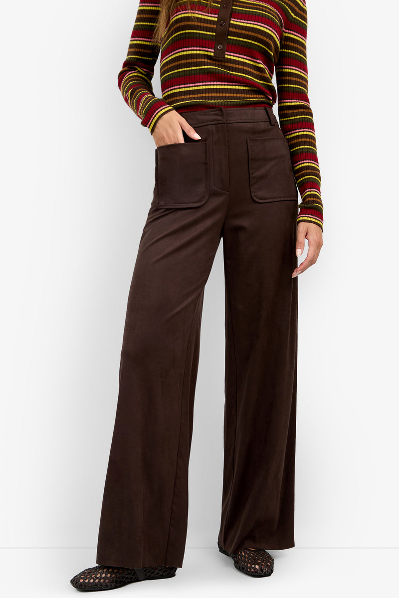 A woman is wearing the ZINN PANT — high-waisted wide-leg brown pants with front pockets — paired with a striped long-sleeve top. She stands facing forward with one hand in her pocket; only her body from neck to feet is visible.