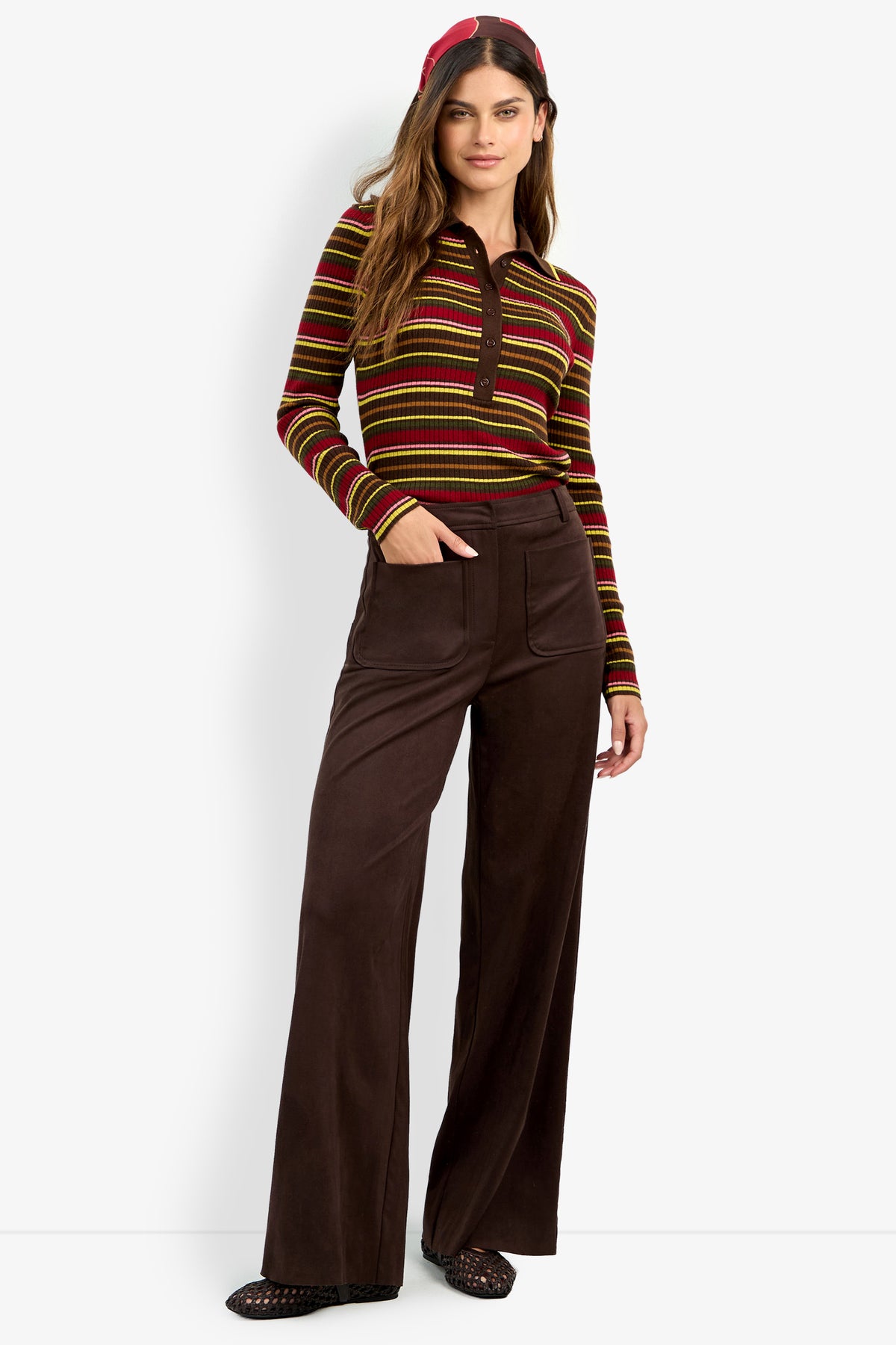 A woman stands against a plain background with one hand in her pocket, wearing the high-waisted brown ZINN PANT, a striped long-sleeve shirt in red, yellow, and brown tones, and a red headband.