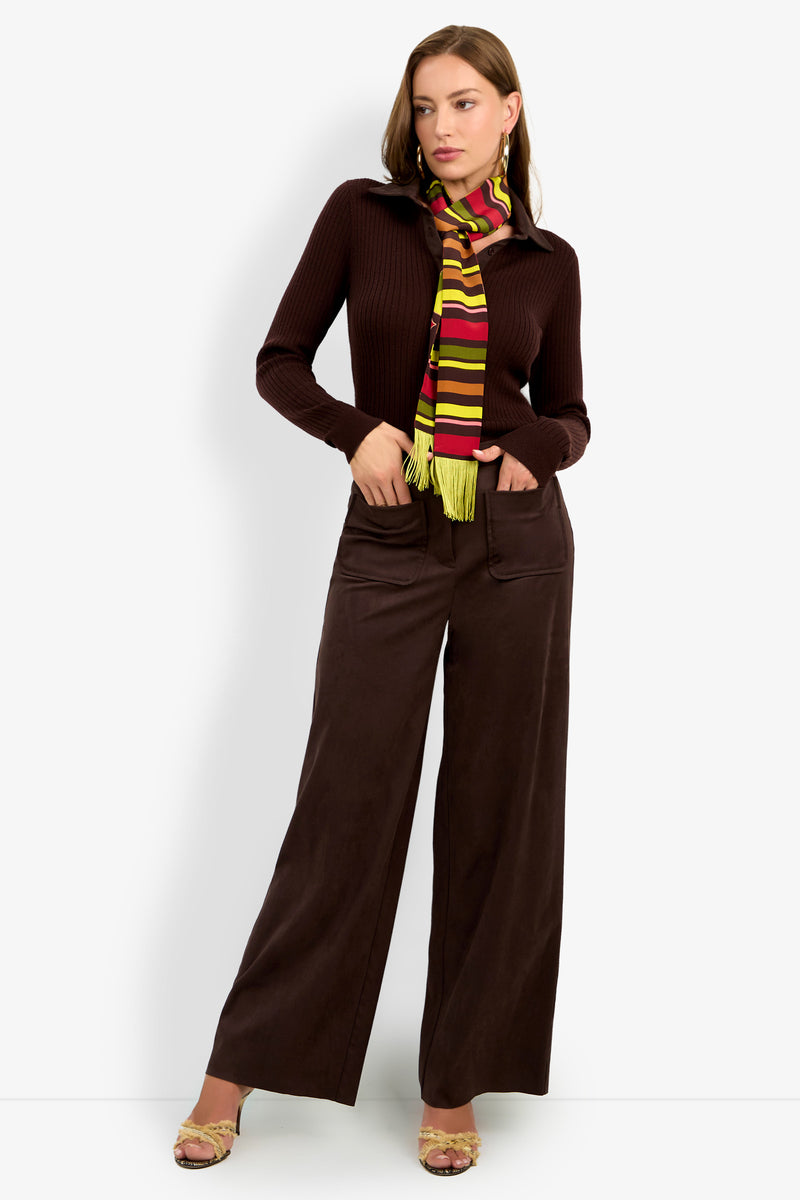 A woman stands against a white background wearing the dark brown ZINN PANT with a matching long-sleeve top and a colorful striped scarf with fringe. She has one hand in her pocket and is wearing open-toe heels.