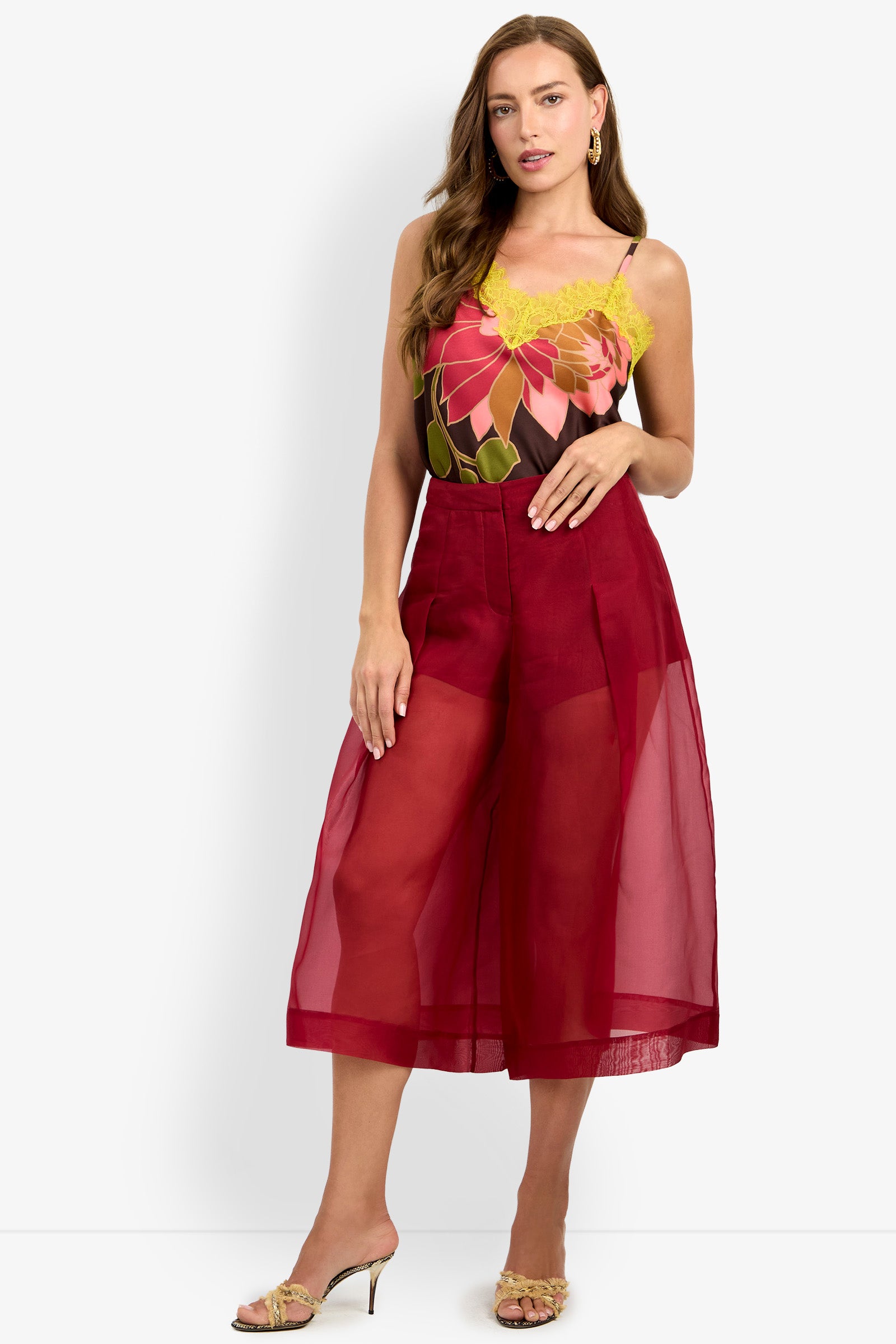 A woman with light brown hair poses against a white background, wearing the SIENA PANT—loose, sheer red culottes—paired with a floral camisole top with thin straps and sandals. She stands with one hand on her hip.