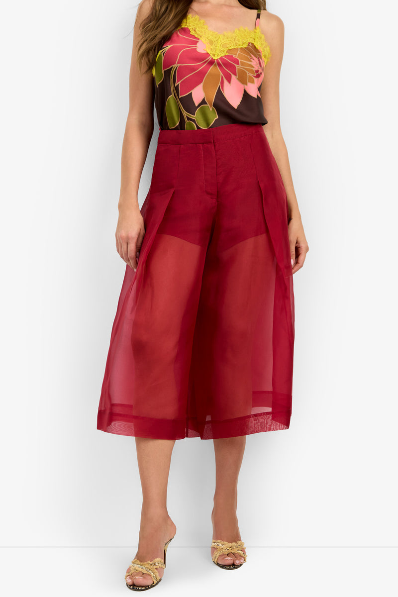 A woman stands against a plain white background, wearing yellow sandals and the SIENA PANT—a pair of sheer red wide-leg culottes—styled with a sleeveless floral top.