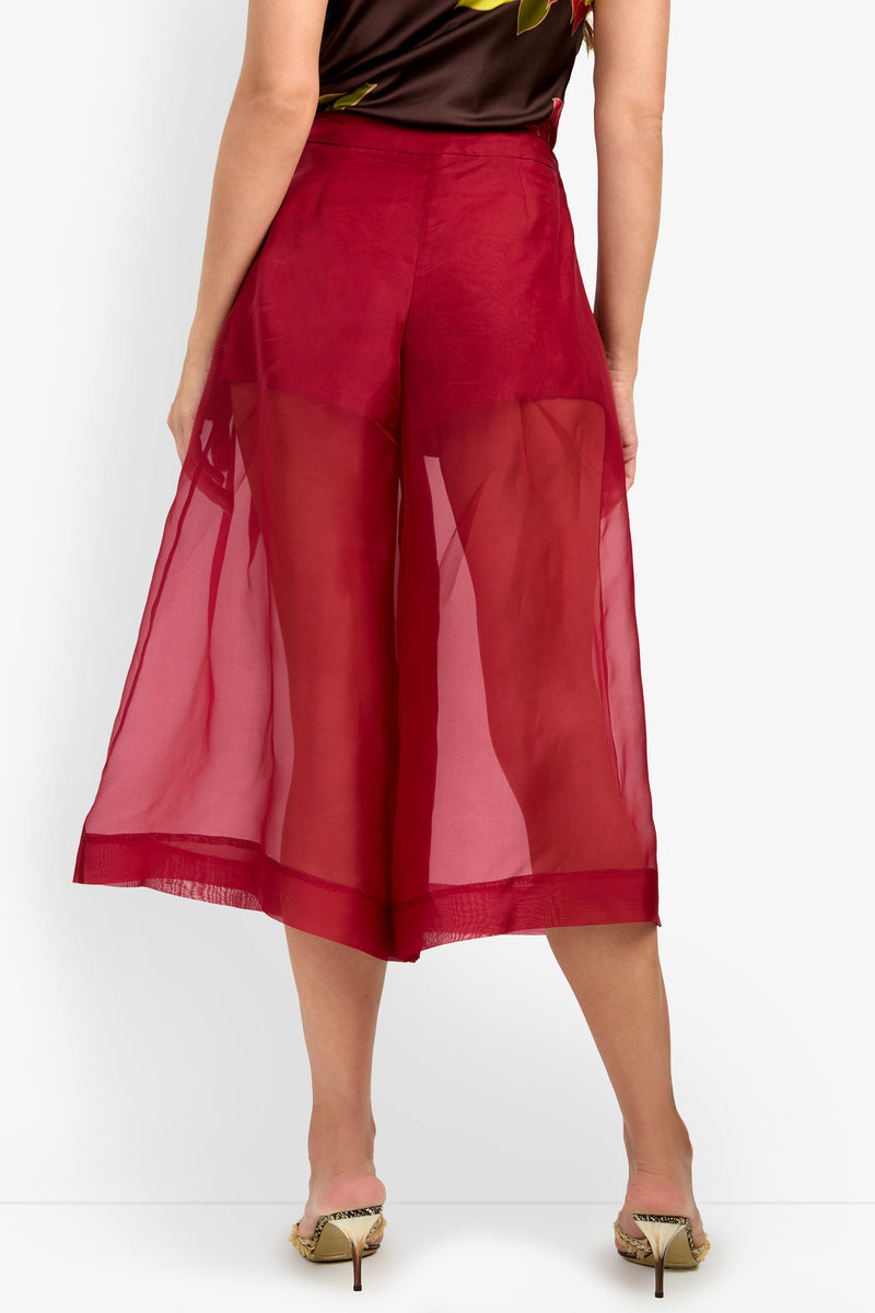 A person wearing the SIENA PANT in sheer red, viewed from the back. The pants are see-through from waist to mid-calf, and the person is standing in heeled sandals on a white background.